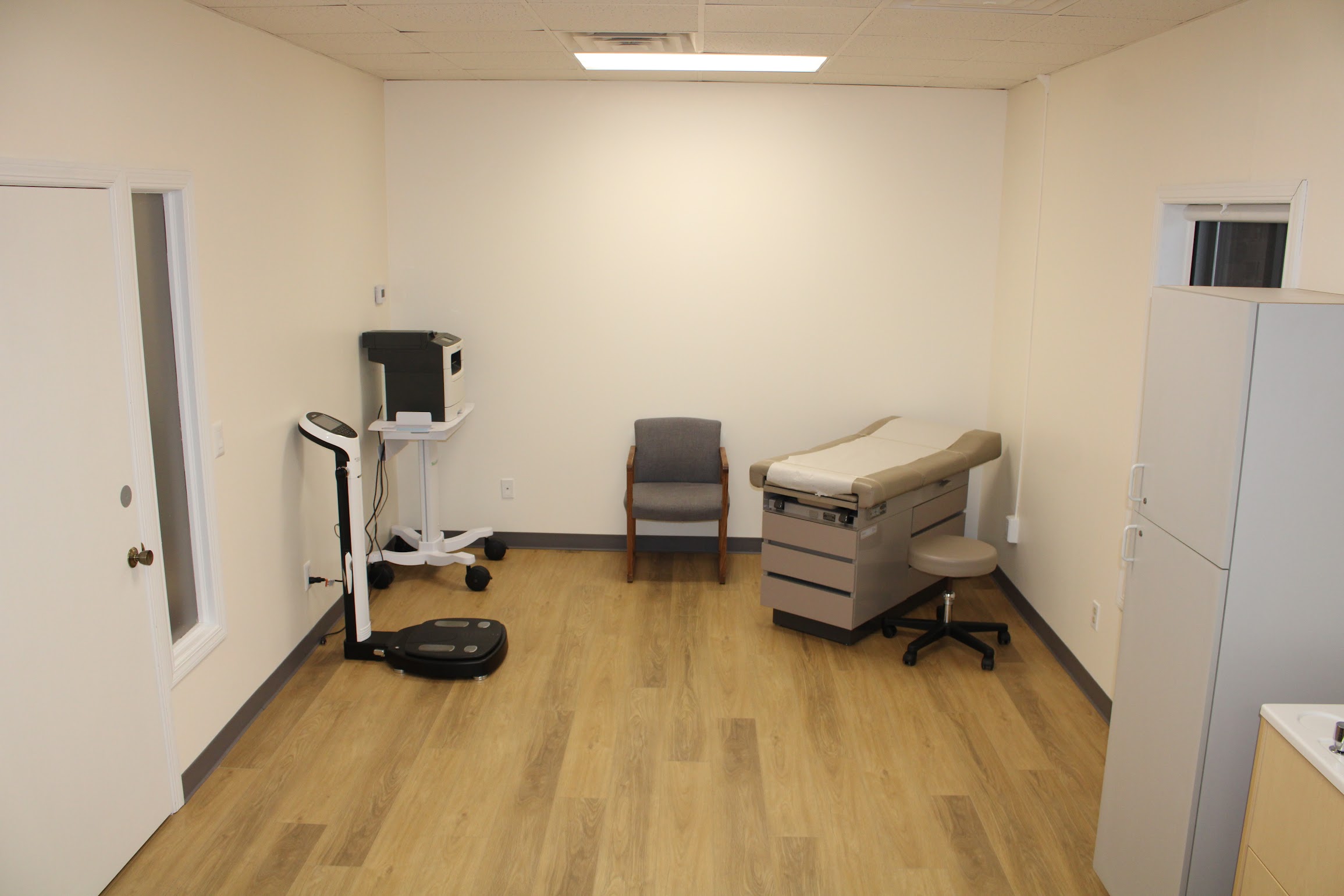 Empty room with medical equipment, chair, and cabinet, hardwood floor, white walls, and ceiling light.