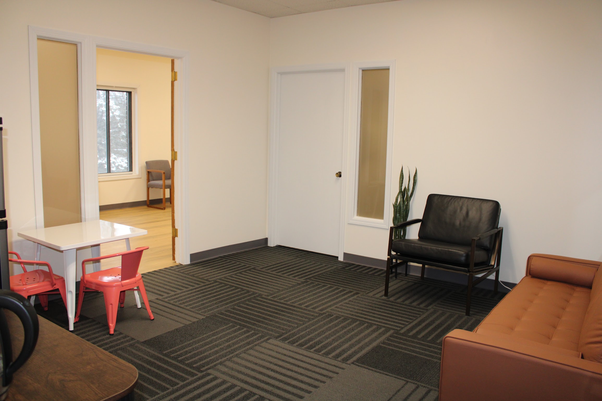 Room with a black chair, brown sofa, white door, and a small plant, with a doorway leading to a space with a chair and window.