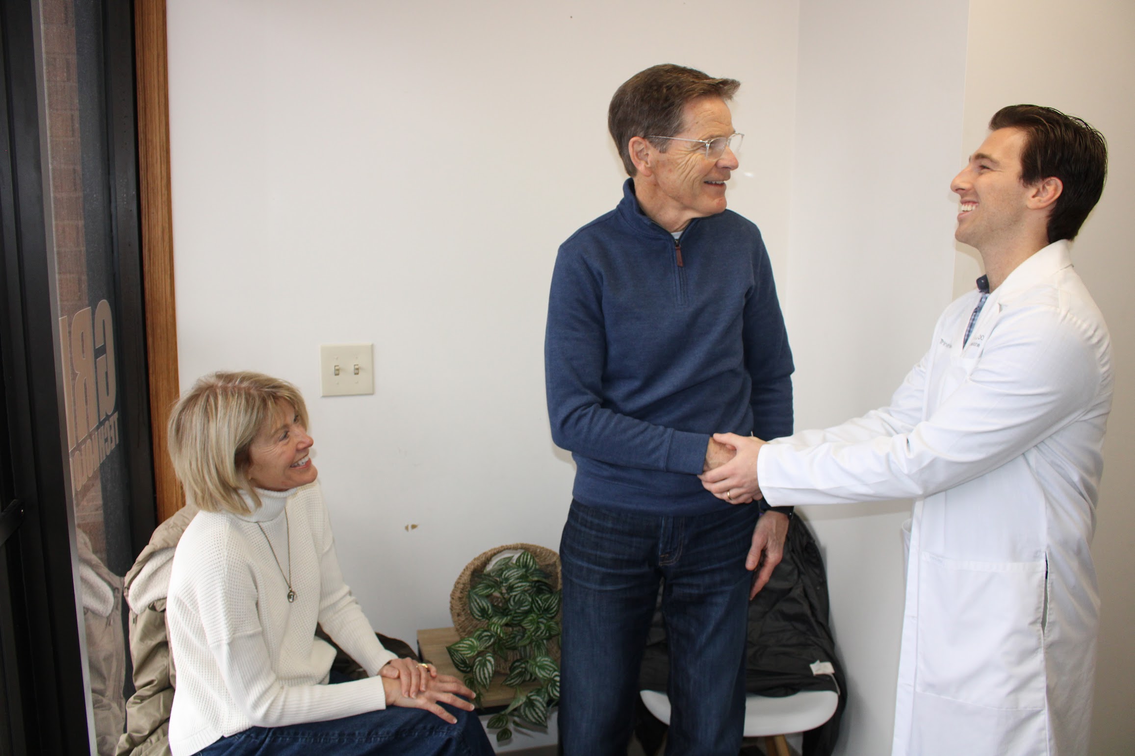 Two men shaking hands, one in a white coat and the other in a blue sweater, with a woman sitting nearby in a room.