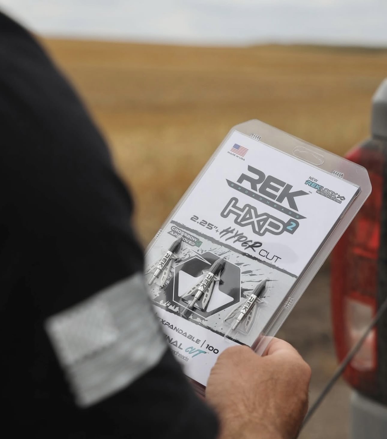 Person holding a smartphone displaying a magazine or brochure with text and images, outdoors with a field in the background.