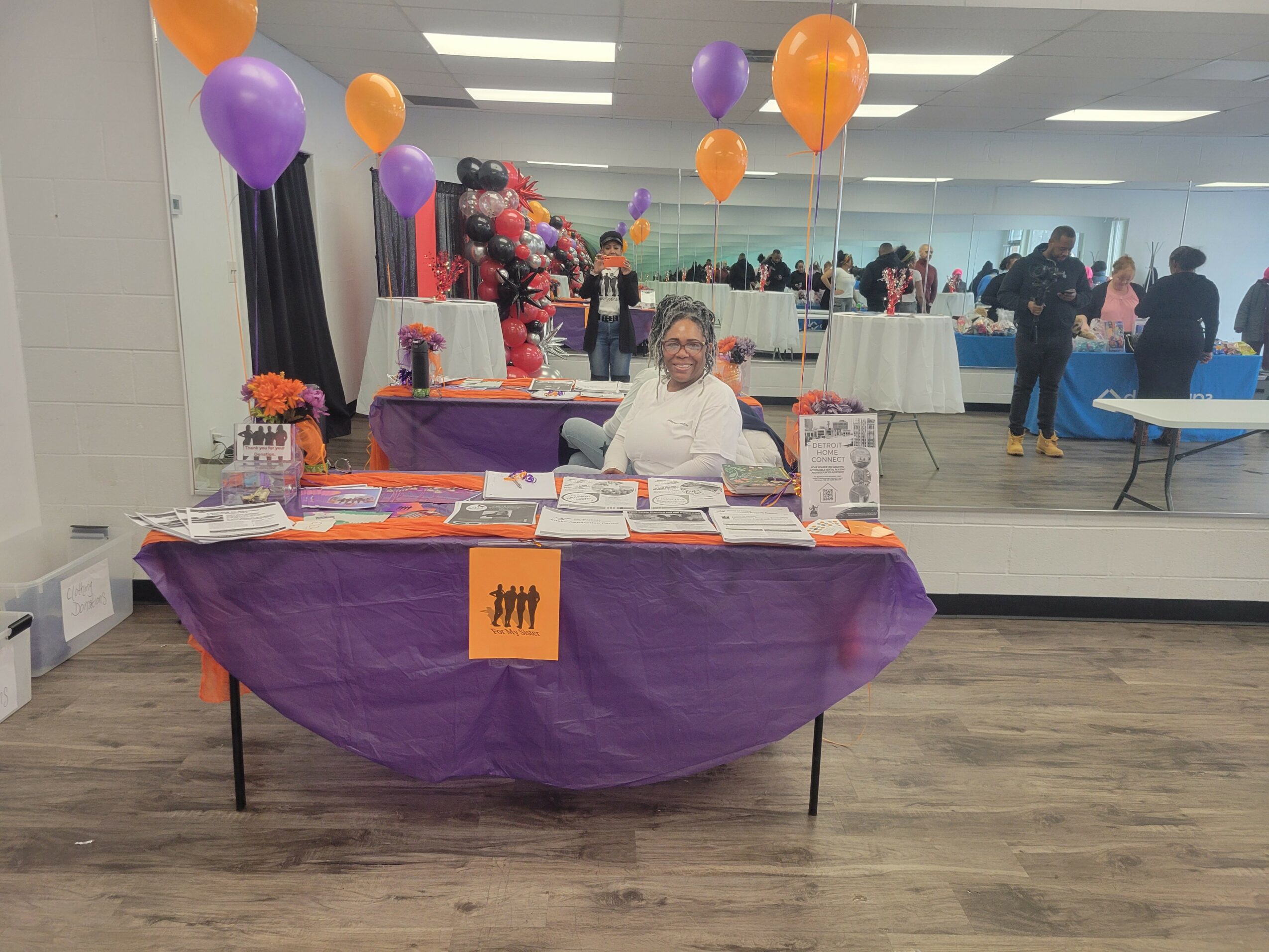 A woman sits at a table decorated with purple and orange balloons, flowers, and informational materials in a large room.
