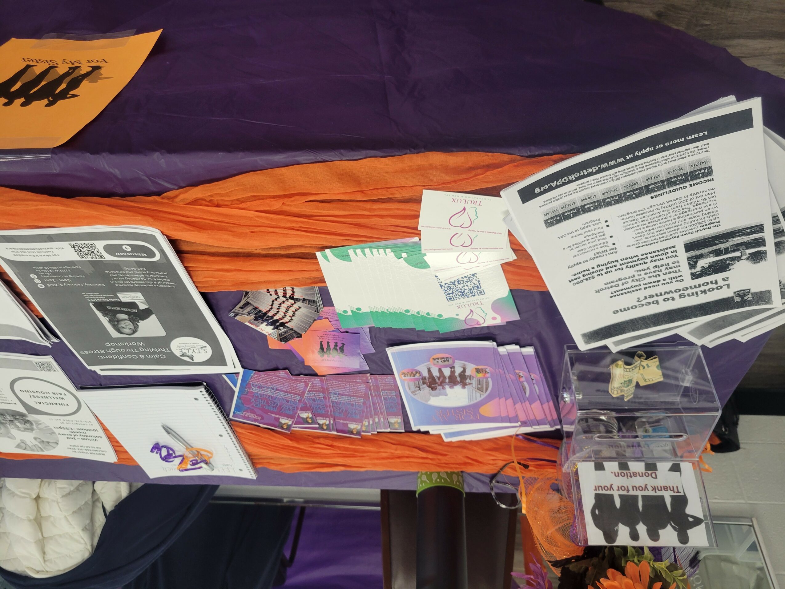 Table with pamphlets, papers, and a plastic container, with a purple and orange fabric background.