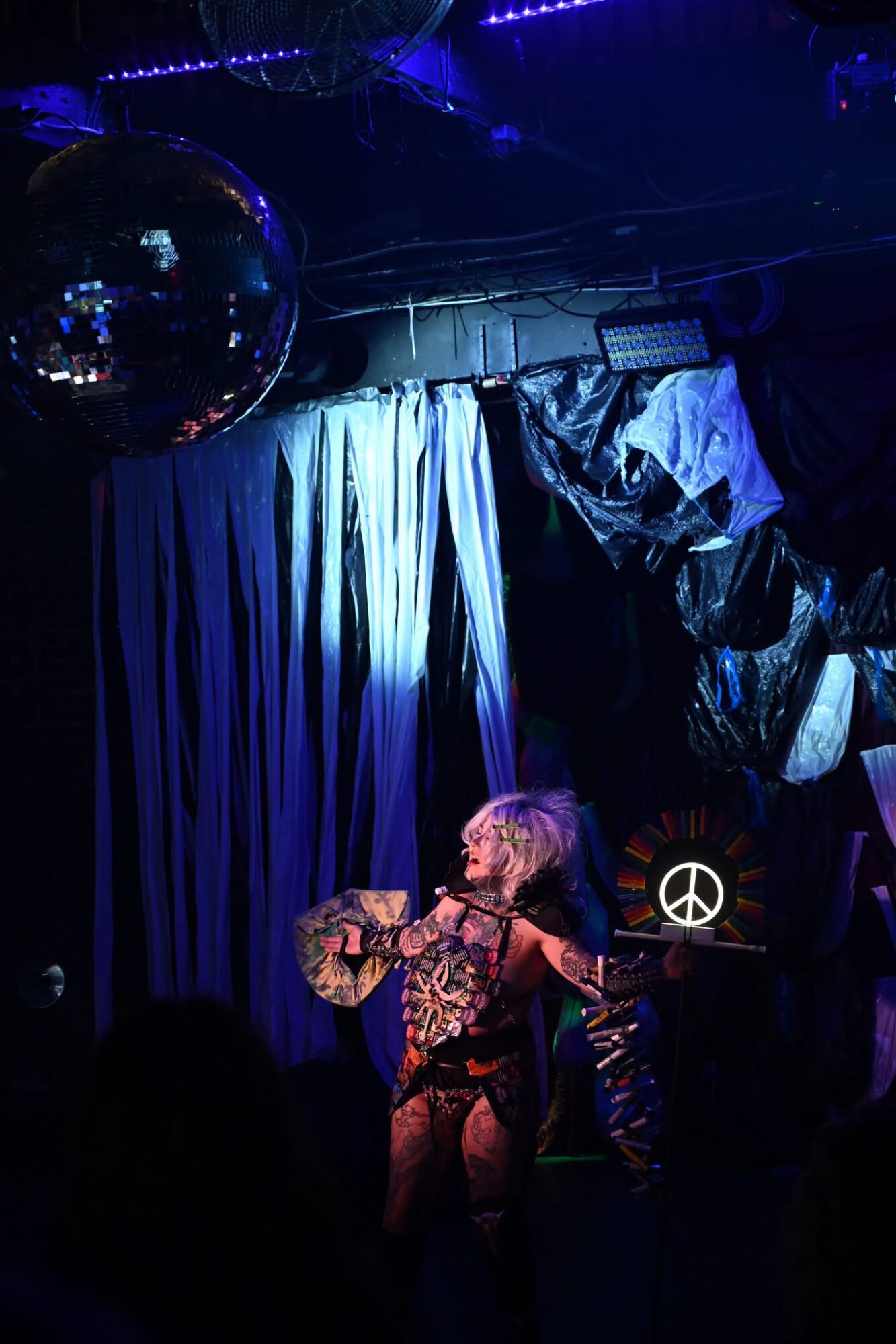 Performer with a guitar on stage, dark background with curtains and disco ball, colorful lighting, audience in foreground.