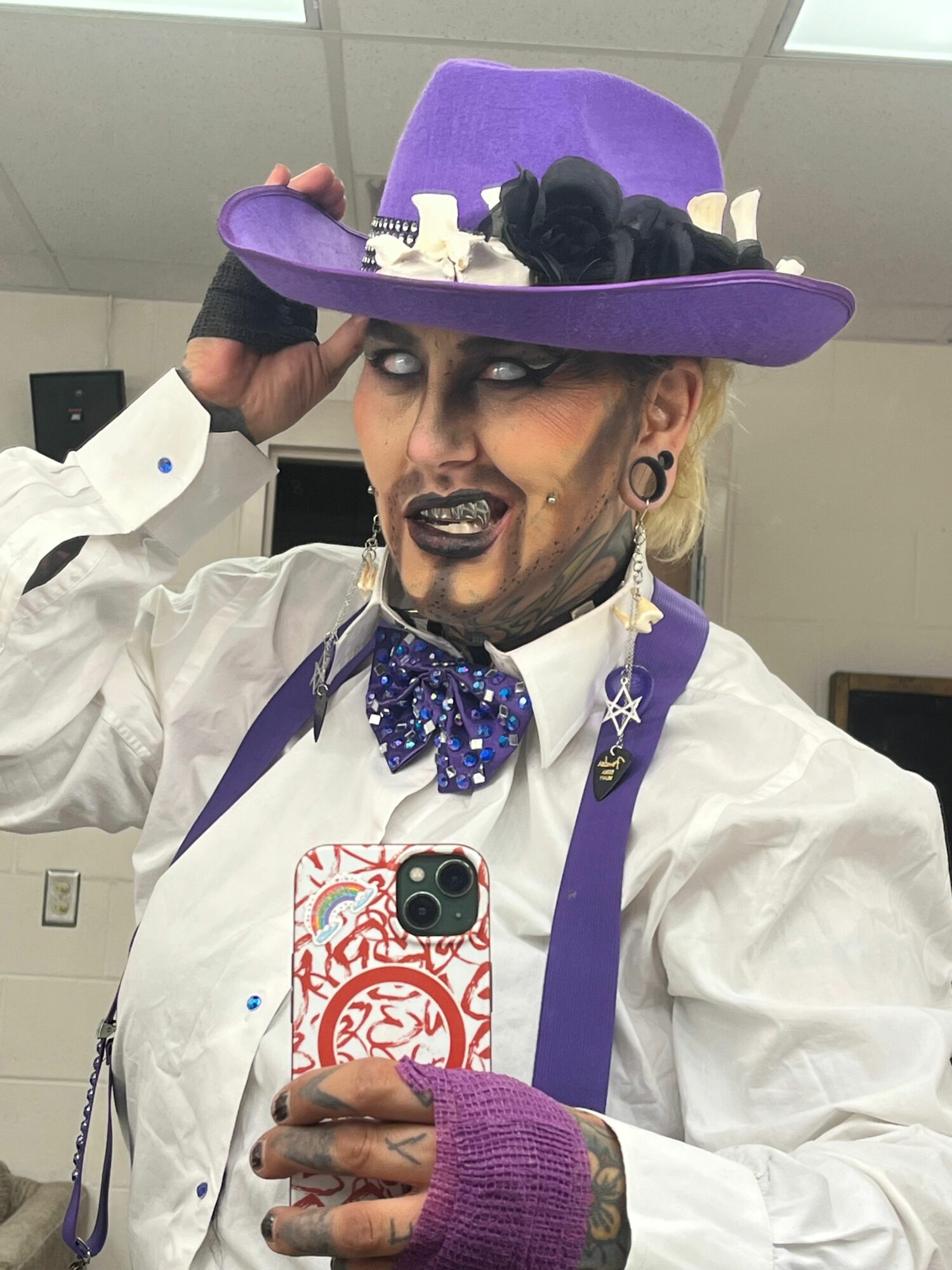 Person wearing a purple hat with black decorations, white shirt, purple bow tie, and purple gloves, smiling and holding a phone.