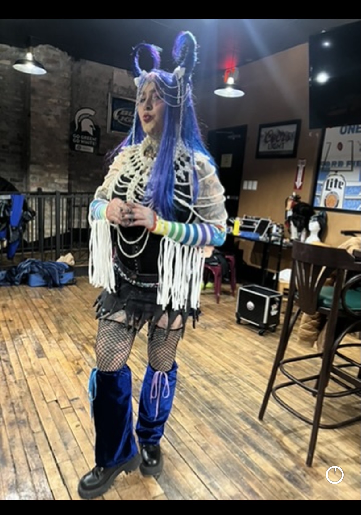 Person in colorful costume with blue hair, horns, and fishnet stockings standing indoors with wooden floor and bar area.