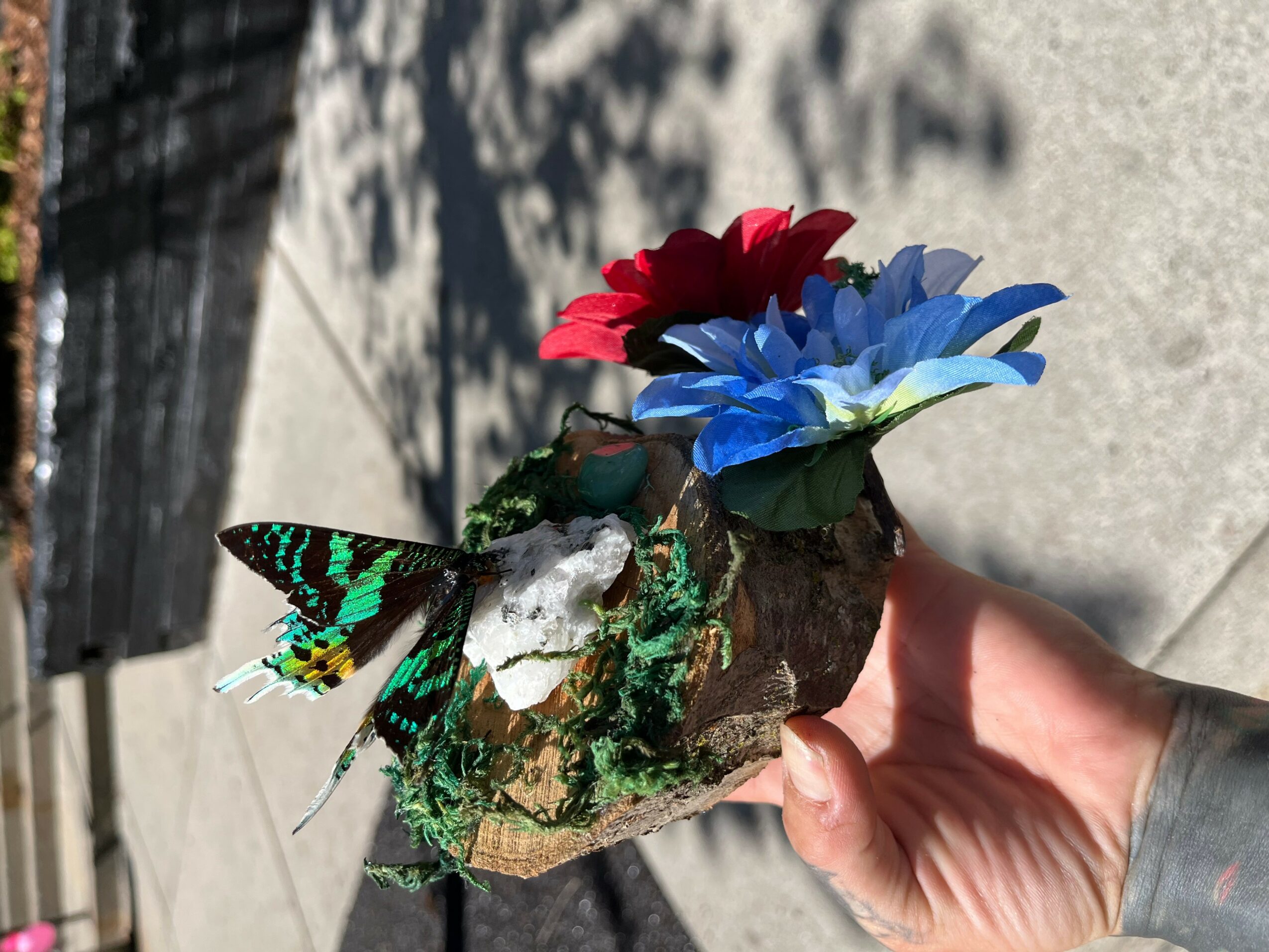 Hand holding a rock with colorful artificial flowers and a butterfly decoration, outdoors on a sunny day.