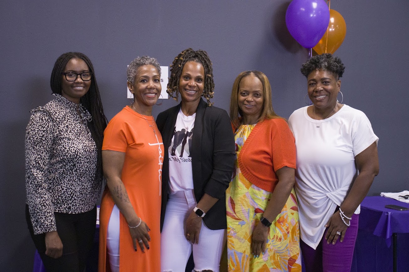 Five women standing together indoors, smiling, with purple and orange balloons in the background.