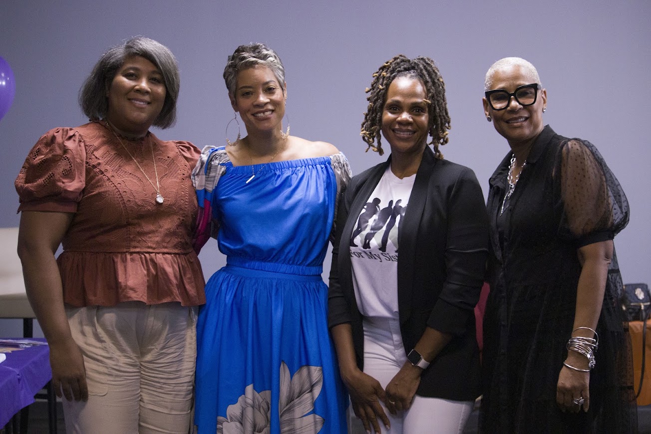 Four women standing together, smiling, in a room with a plain background.