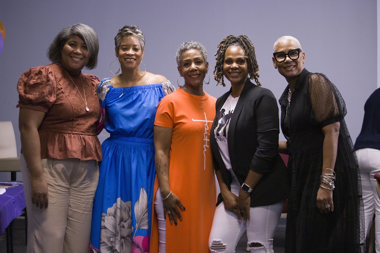 Five women standing together, smiling, in a room with a plain background.