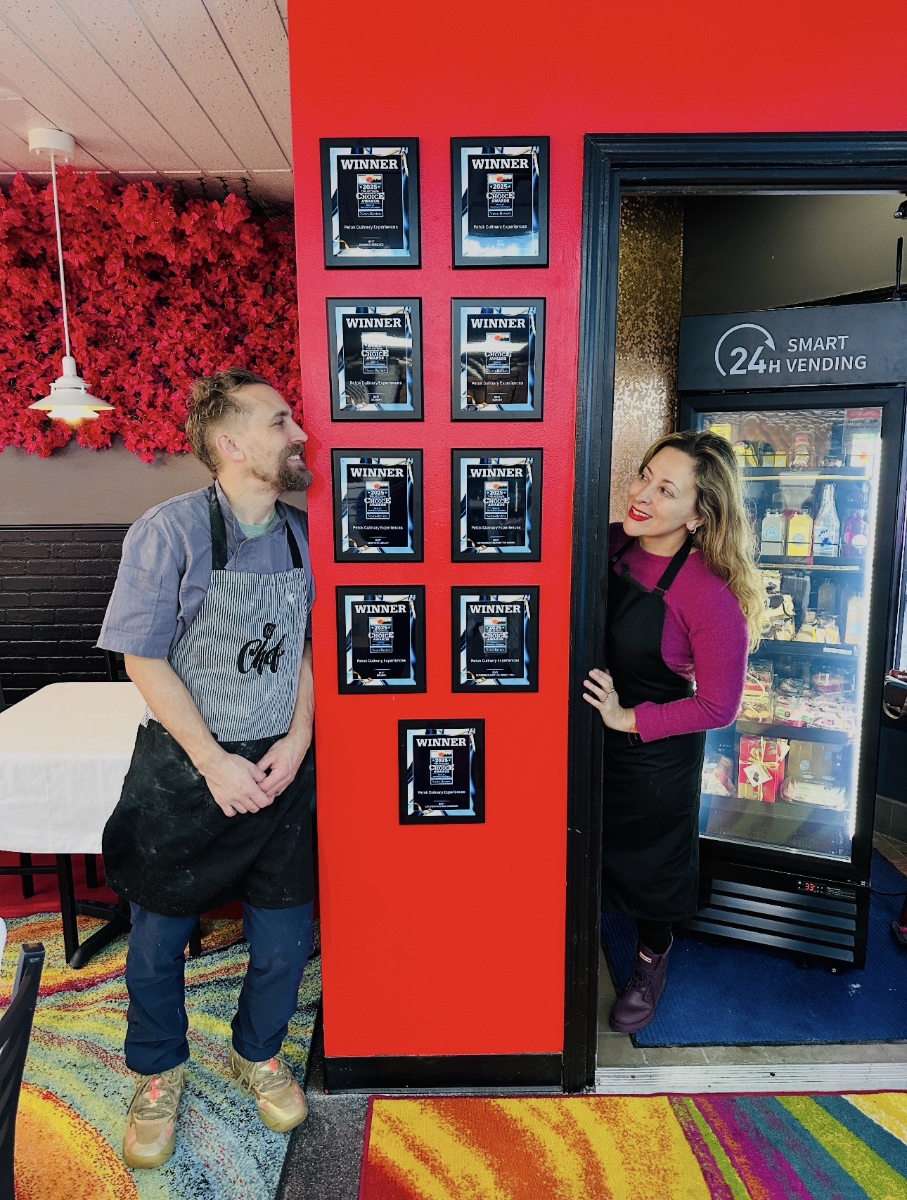 Two women stand near a vending machine and a display with multiple screens, in a colorful room with a red wall and rainbow carpet.