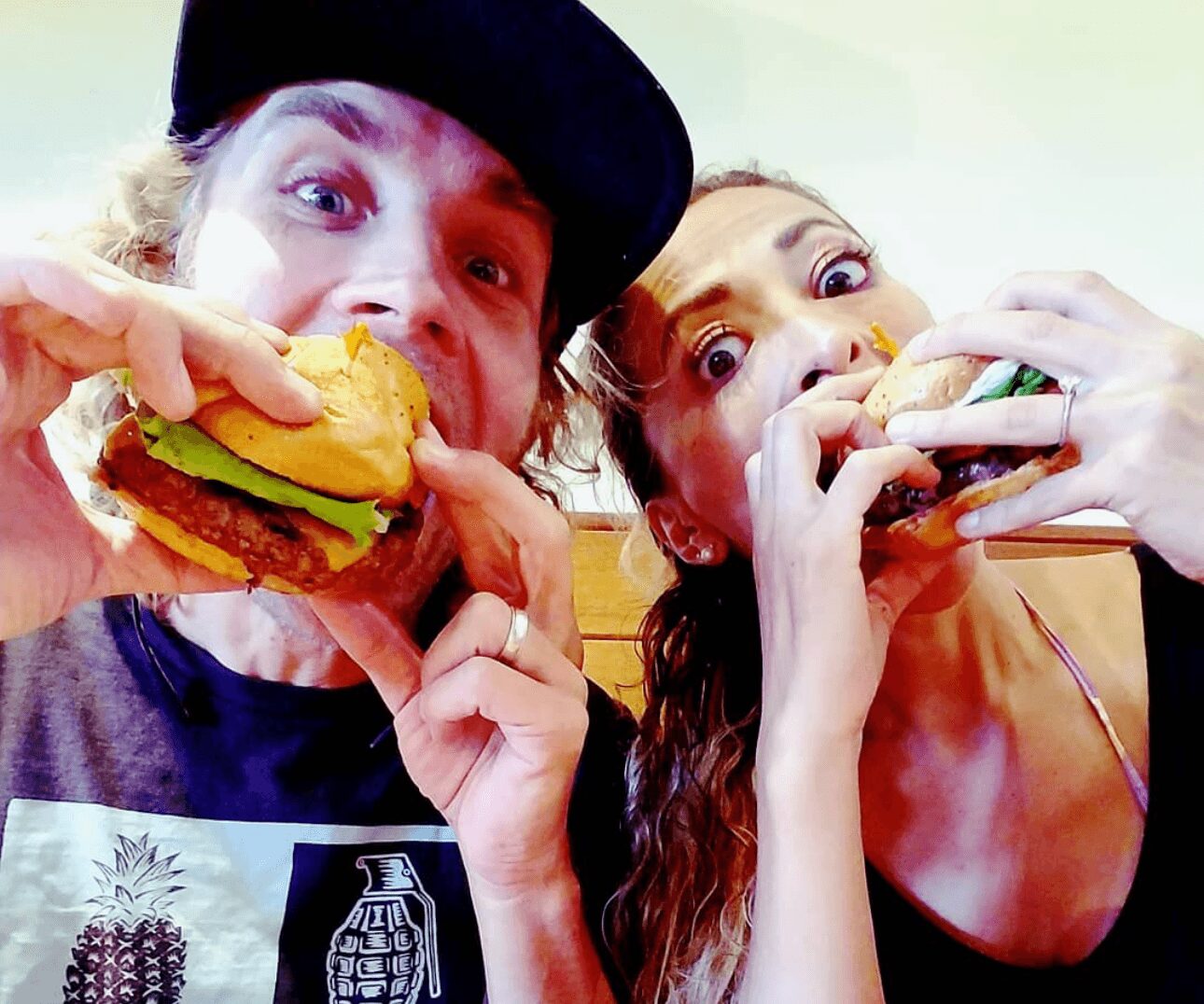 Two people eating large sandwiches, close-up, smiling, with bright expressions, in an indoor setting.