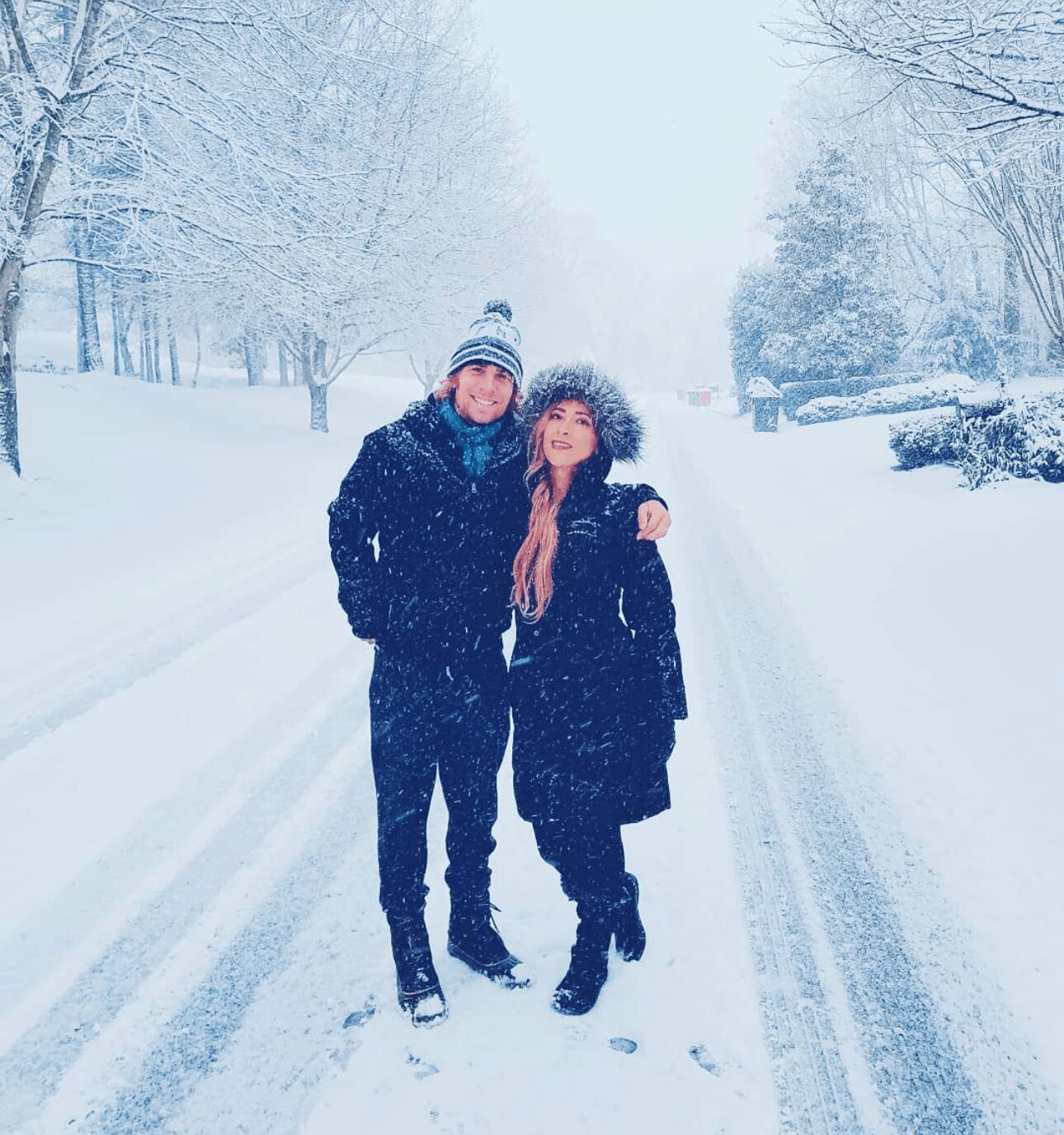 Two people standing on a snowy road with trees on both sides, wearing winter clothing and hats, in a snowy landscape.