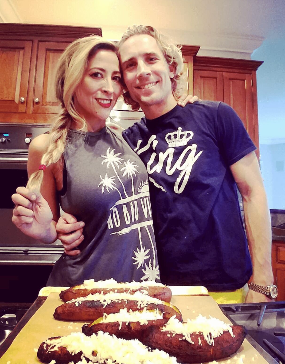Two people smiling in a kitchen, standing behind a cutting board with bread topped with cheese, woman with blonde hair, man with curly hair