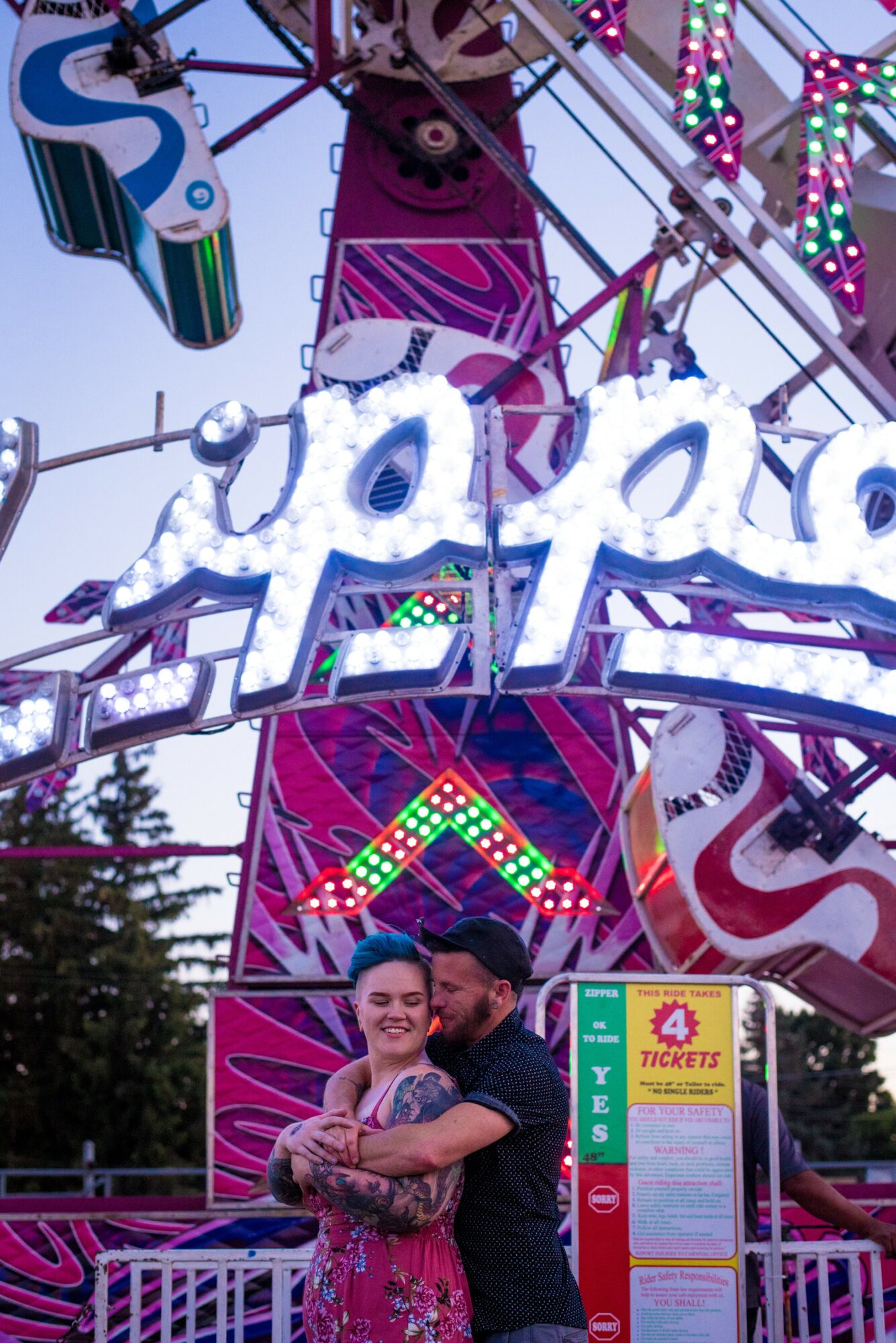 Two people smiling and hugging in front of a colorful amusement park ride with bright lights.