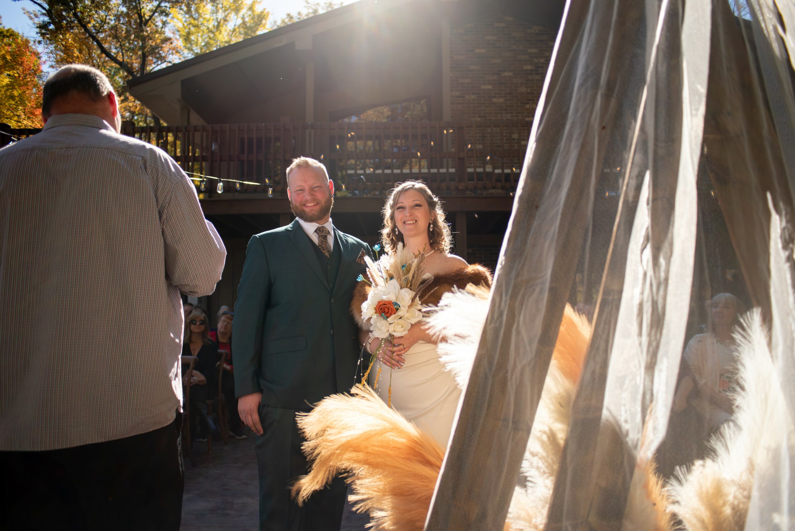 A bride holding a bouquet stands next to a groom outdoors, with sunlight shining behind them, and a tent partially visible.