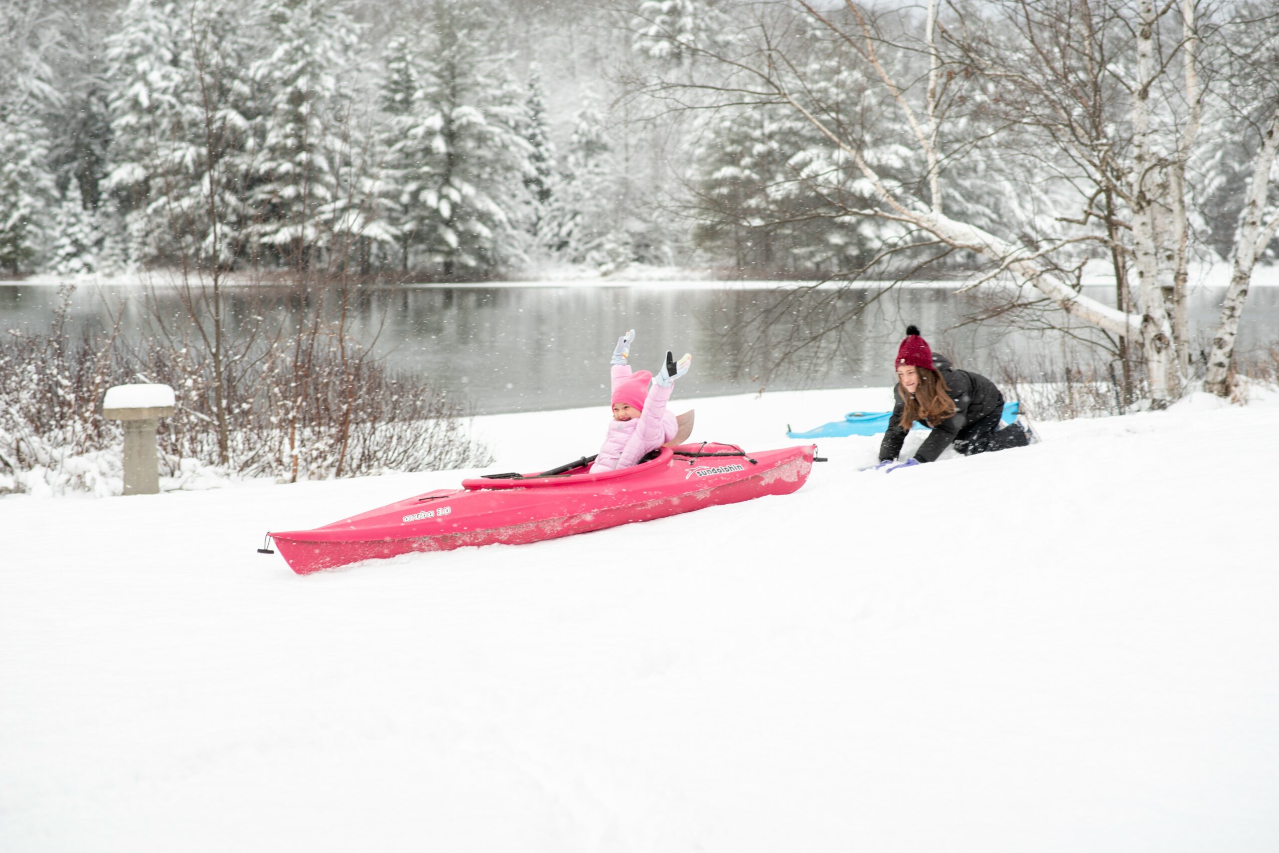 Person in pink jacket and hat in a red kayak on snow-covered ground near a river, with another person nearby, winter scene.