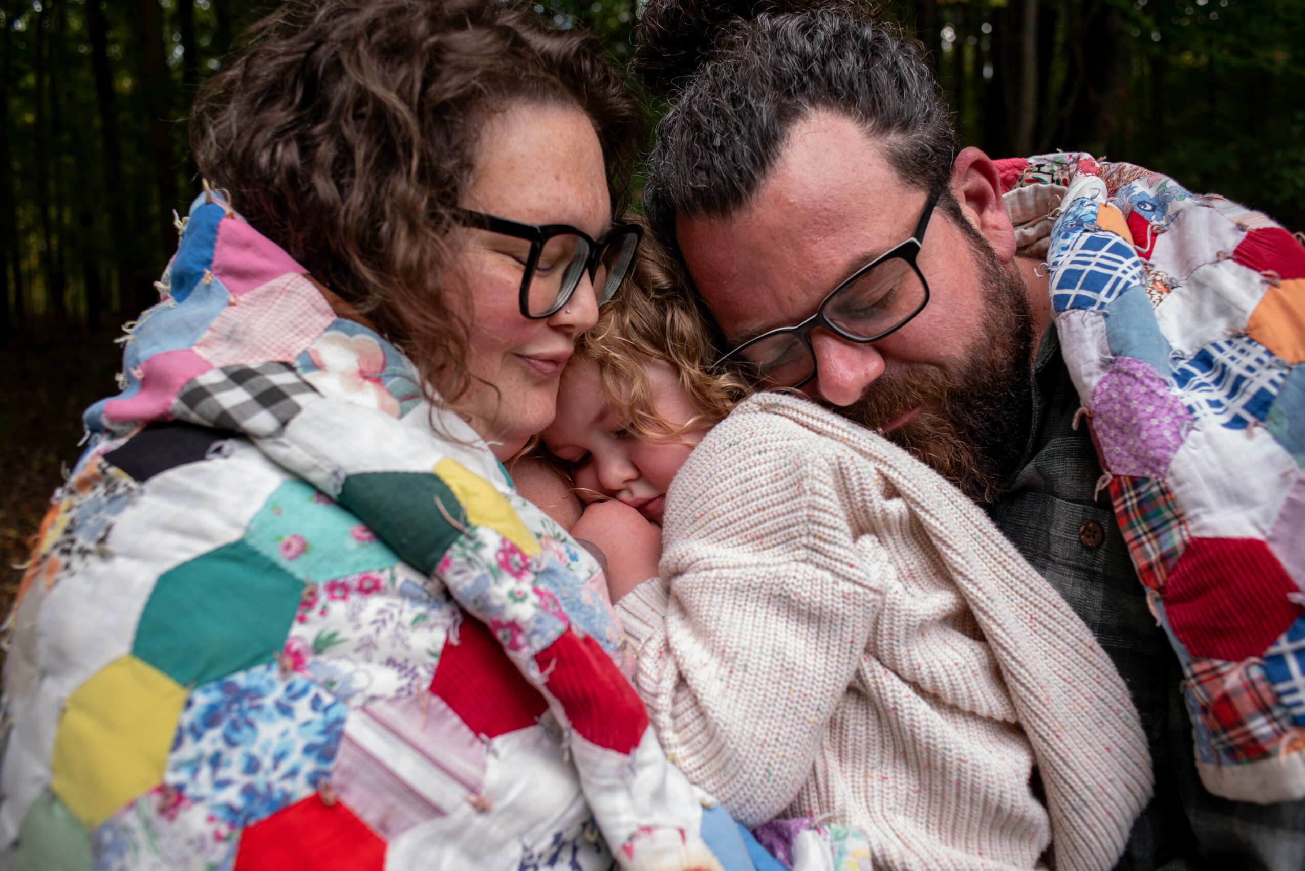 Three people, two adults and a child, close together outdoors, with the child resting on the adults' shoulders, wrapped in a colorful quilt.