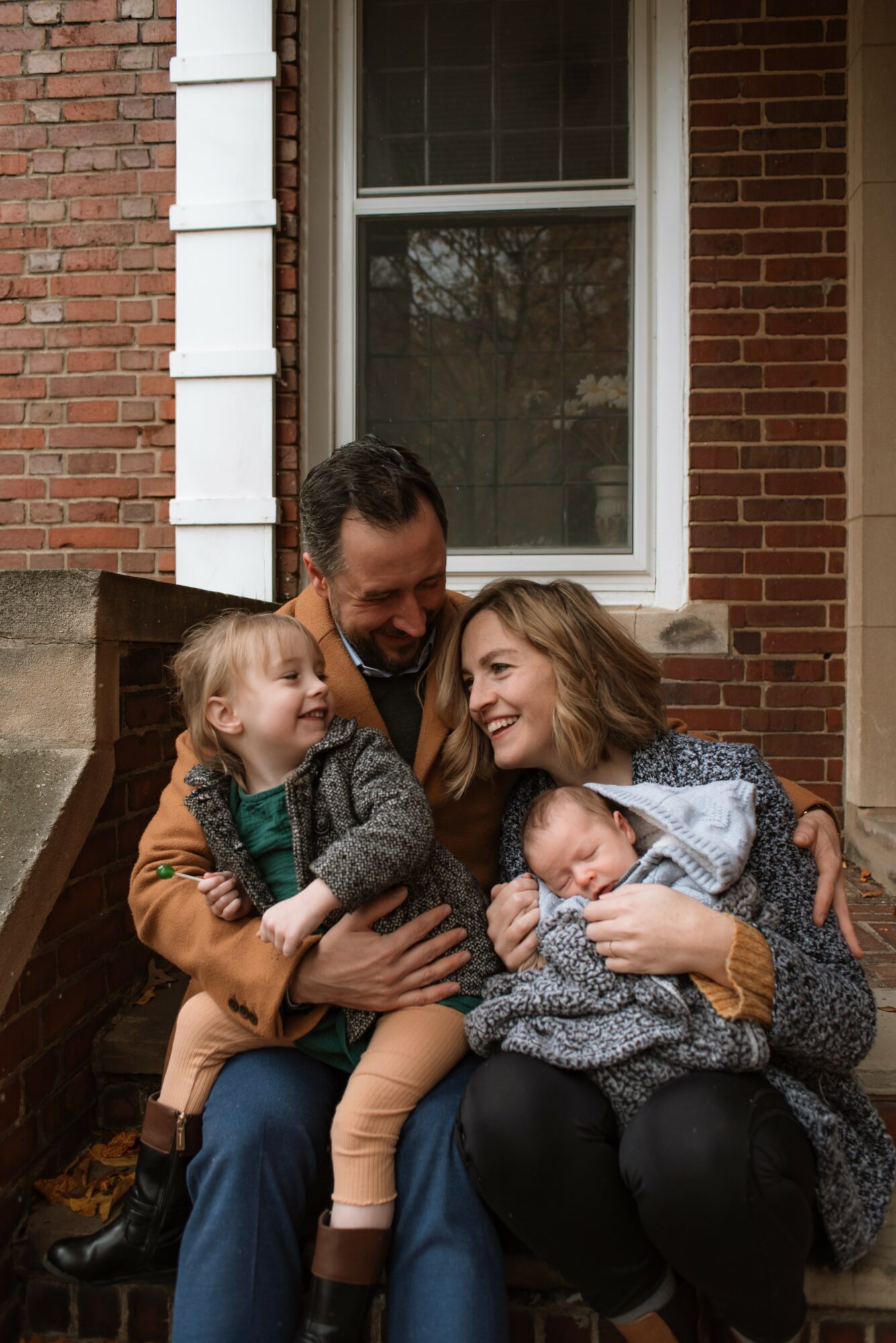 Family of four sitting outside a brick house, smiling and hugging each other, with a window behind them.