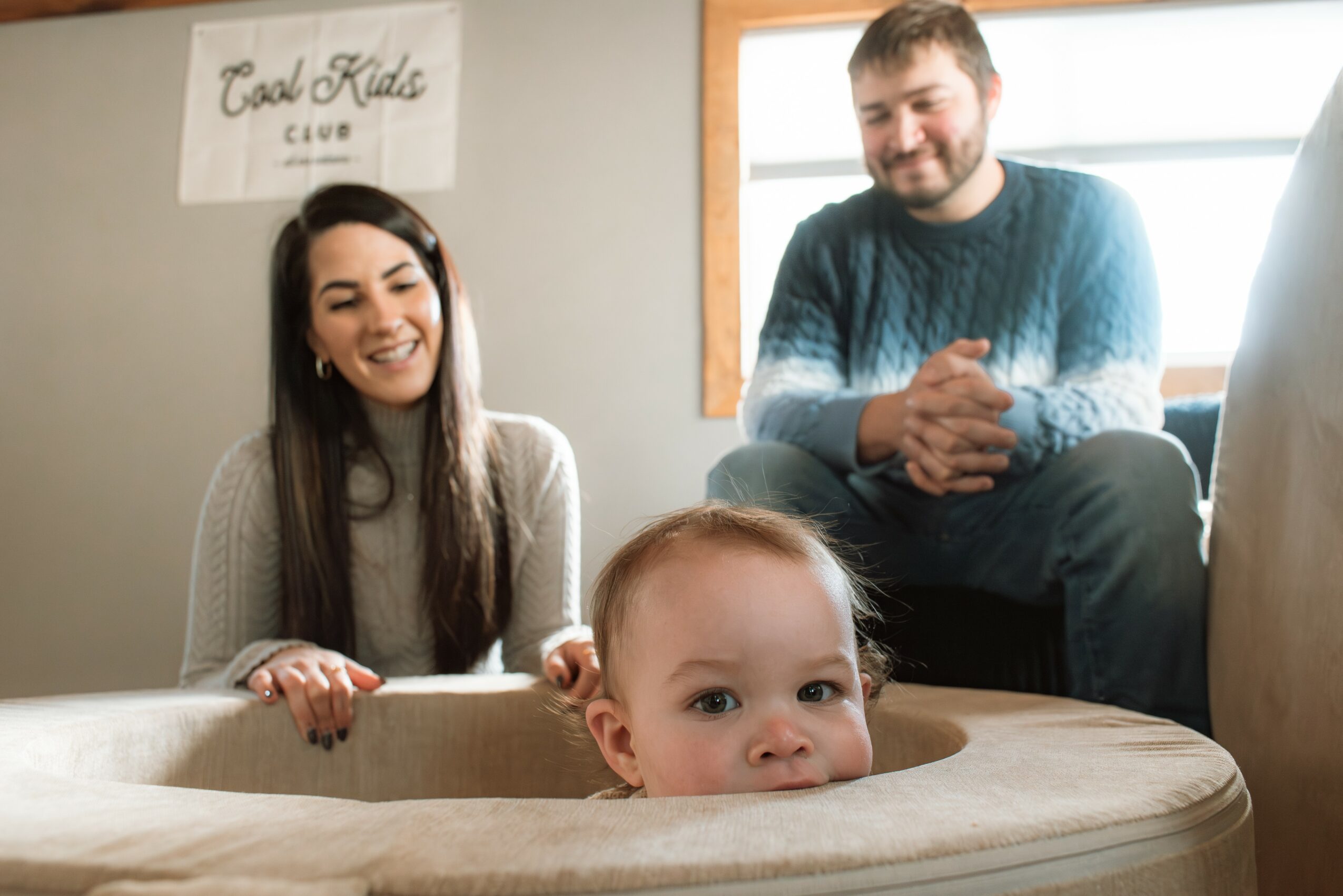 Young child with head in a wooden play area, smiling, with two adults smiling in the background in a room.