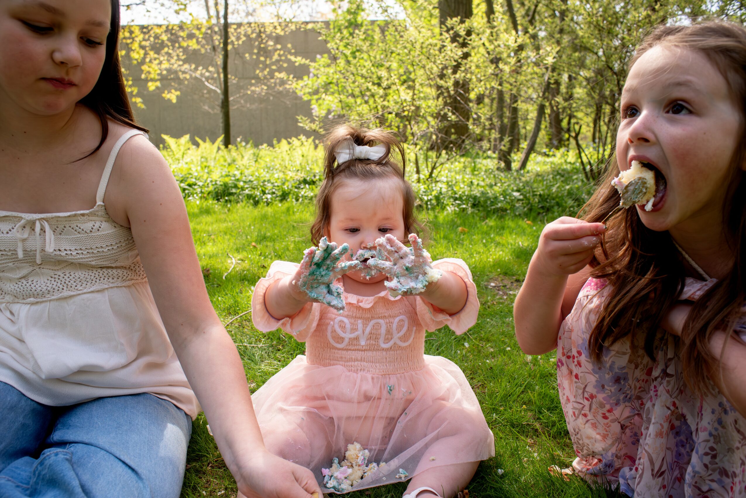Three young girls outdoors, one eating a snack, one holding a cake, and one reaching out, with trees in background.