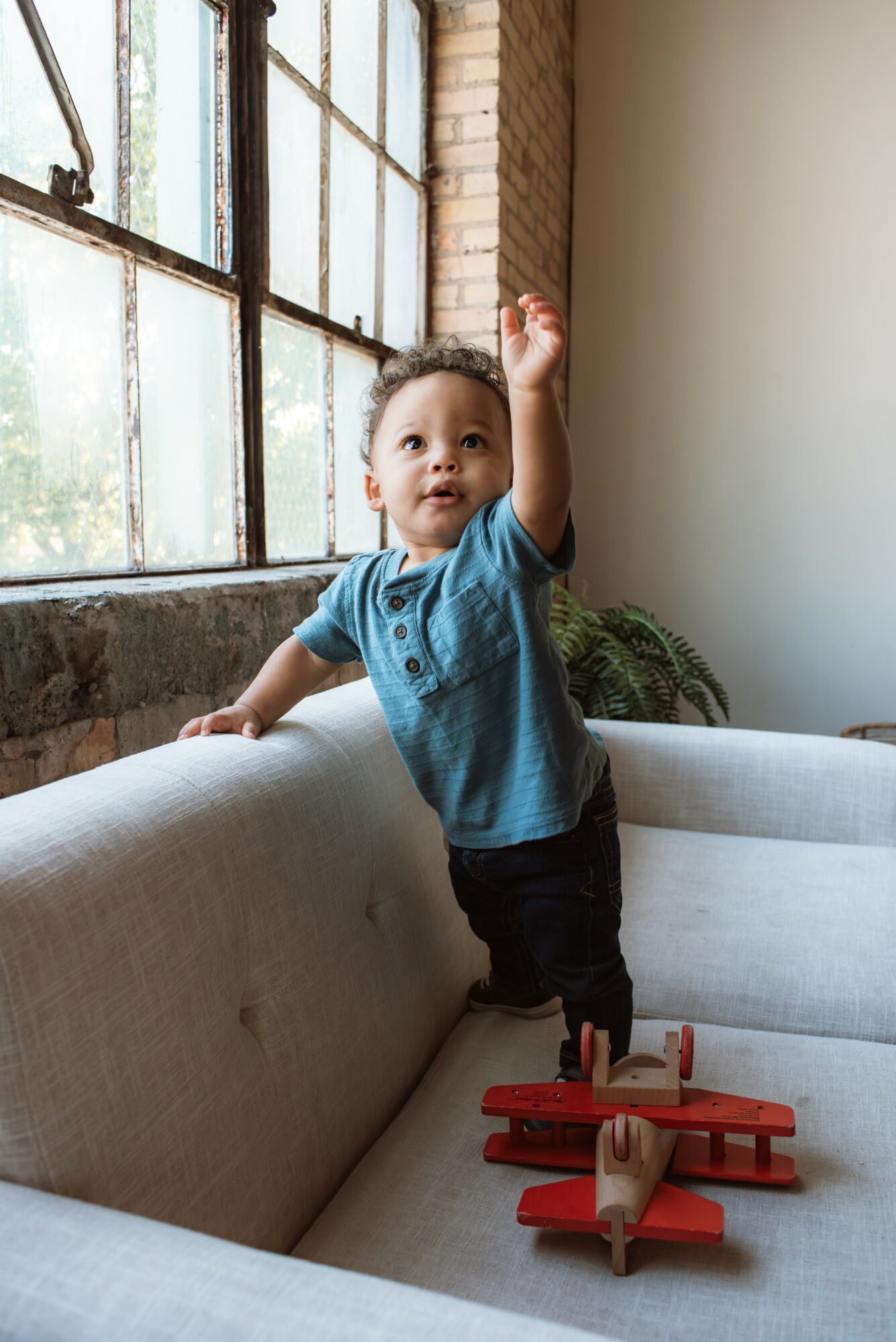 Child standing on sofa, reaching up with one hand, near large window with brick wall, toys on sofa, plant nearby.