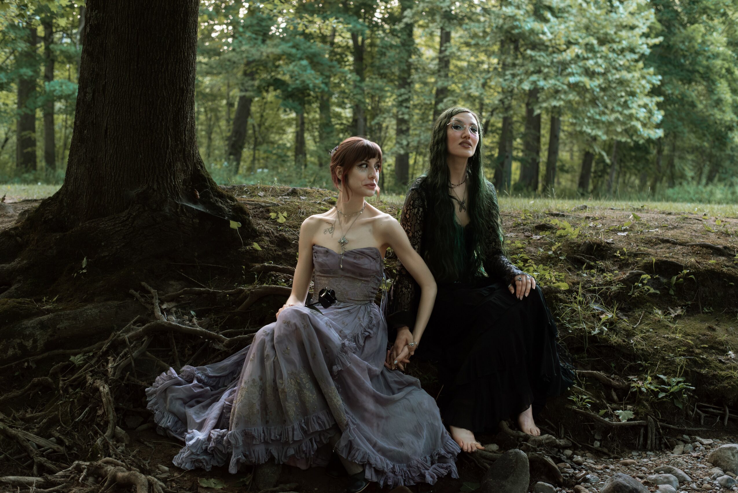 Two women sit on the ground in a forest, one in a gray dress and the other in a black dress, surrounded by trees.