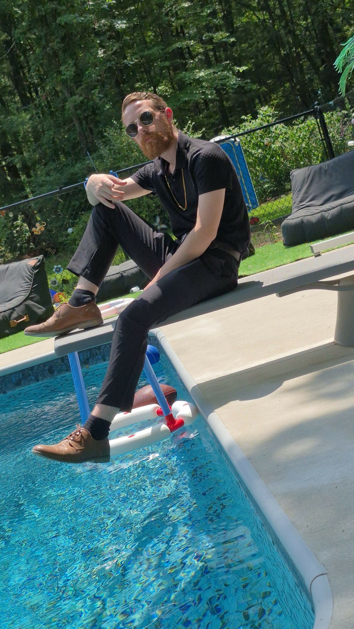 Person crouching by a swimming pool, holding a pool cleaning tool, outdoors with trees in the background.