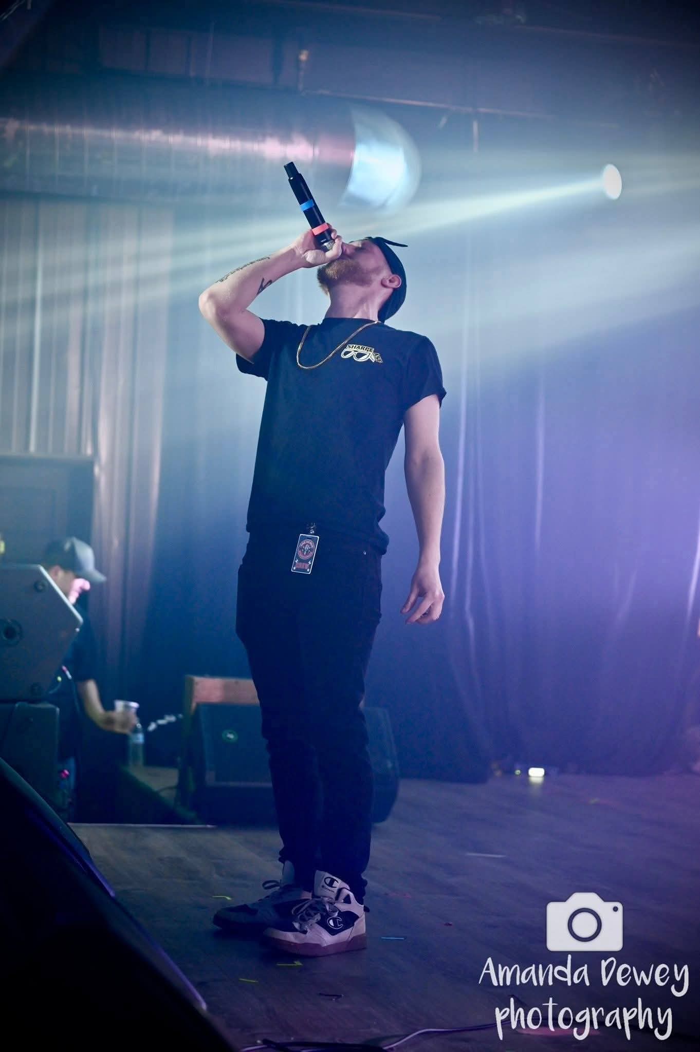 Person drinking from a bottle on stage with colorful lighting and fog effects, wearing a black shirt and sneakers.