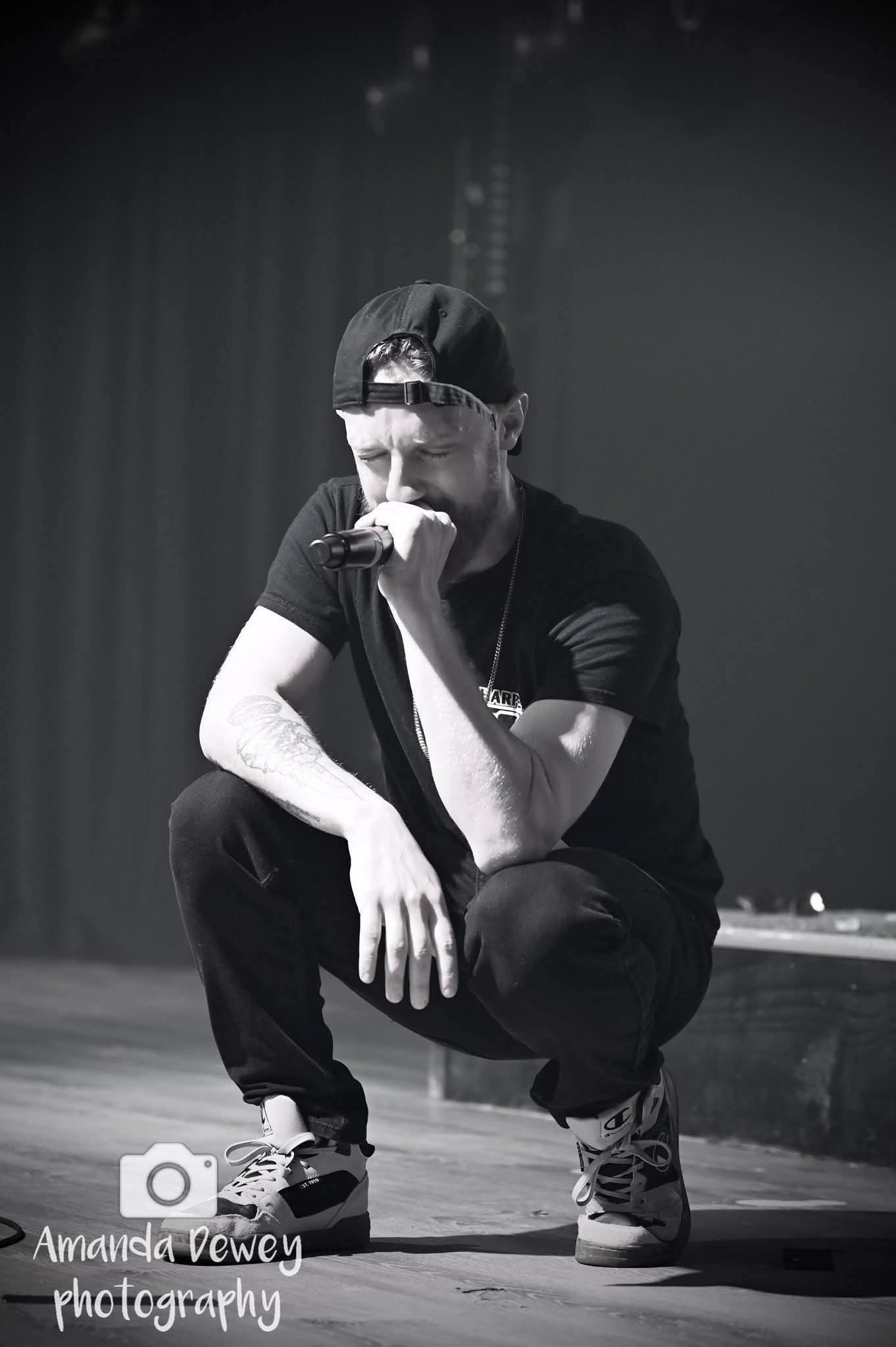 Person crouching on stage, wearing a cap, black t-shirt, and sneakers, with a hand near face, in a performance setting.