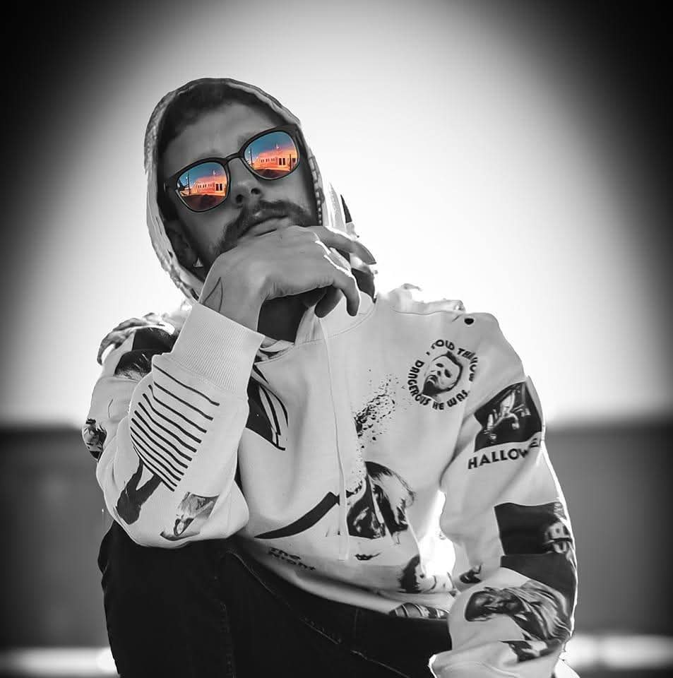 Person wearing sunglasses and a patterned hoodie, resting chin on hand, with a blurred background.