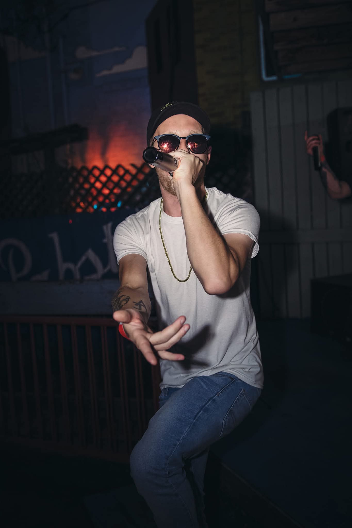 Person wearing sunglasses and a white shirt, holding a microphone, performing on stage in a dark setting.