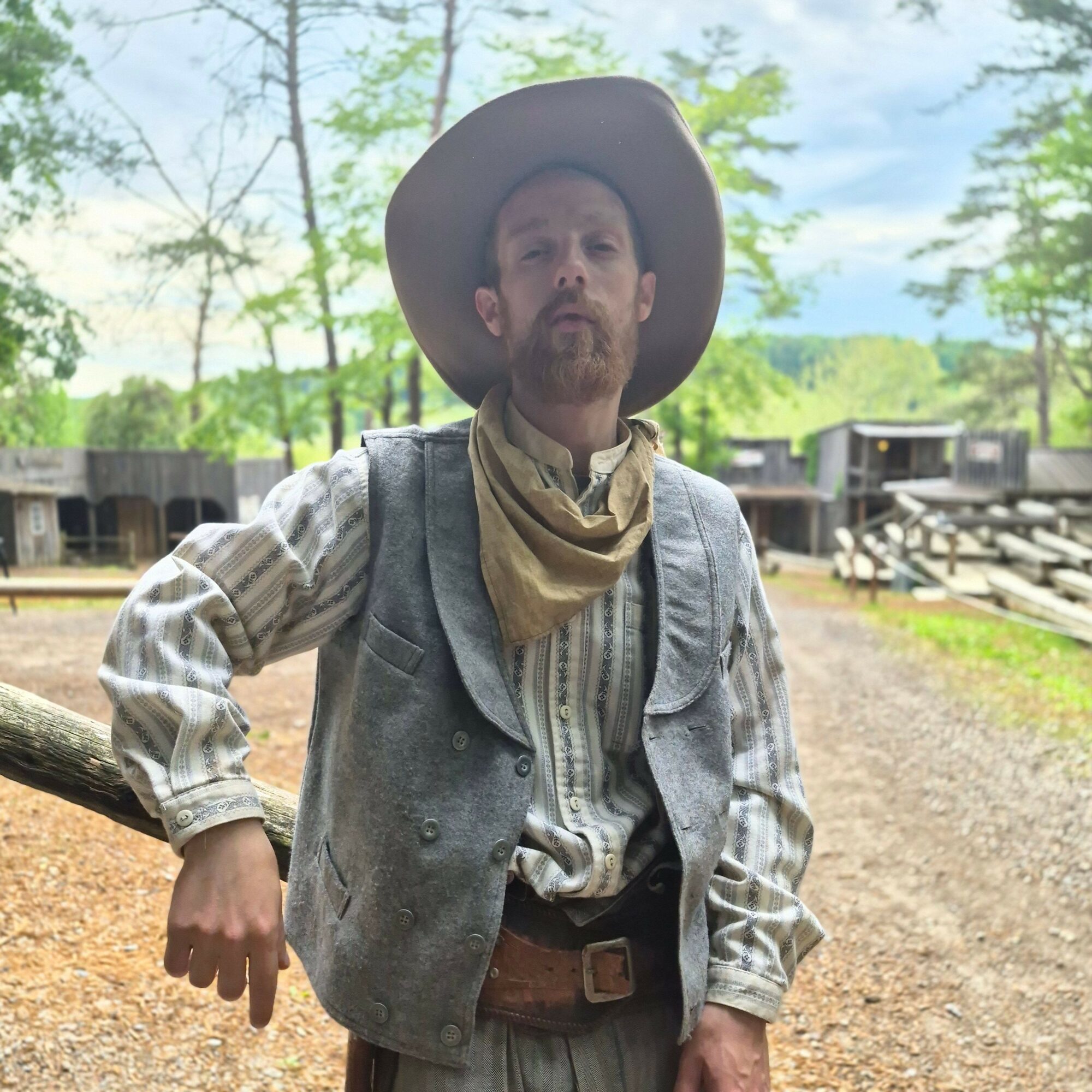Man dressed as a cowboy with a hat, scarf, and vest outdoors, pointing with his right hand.