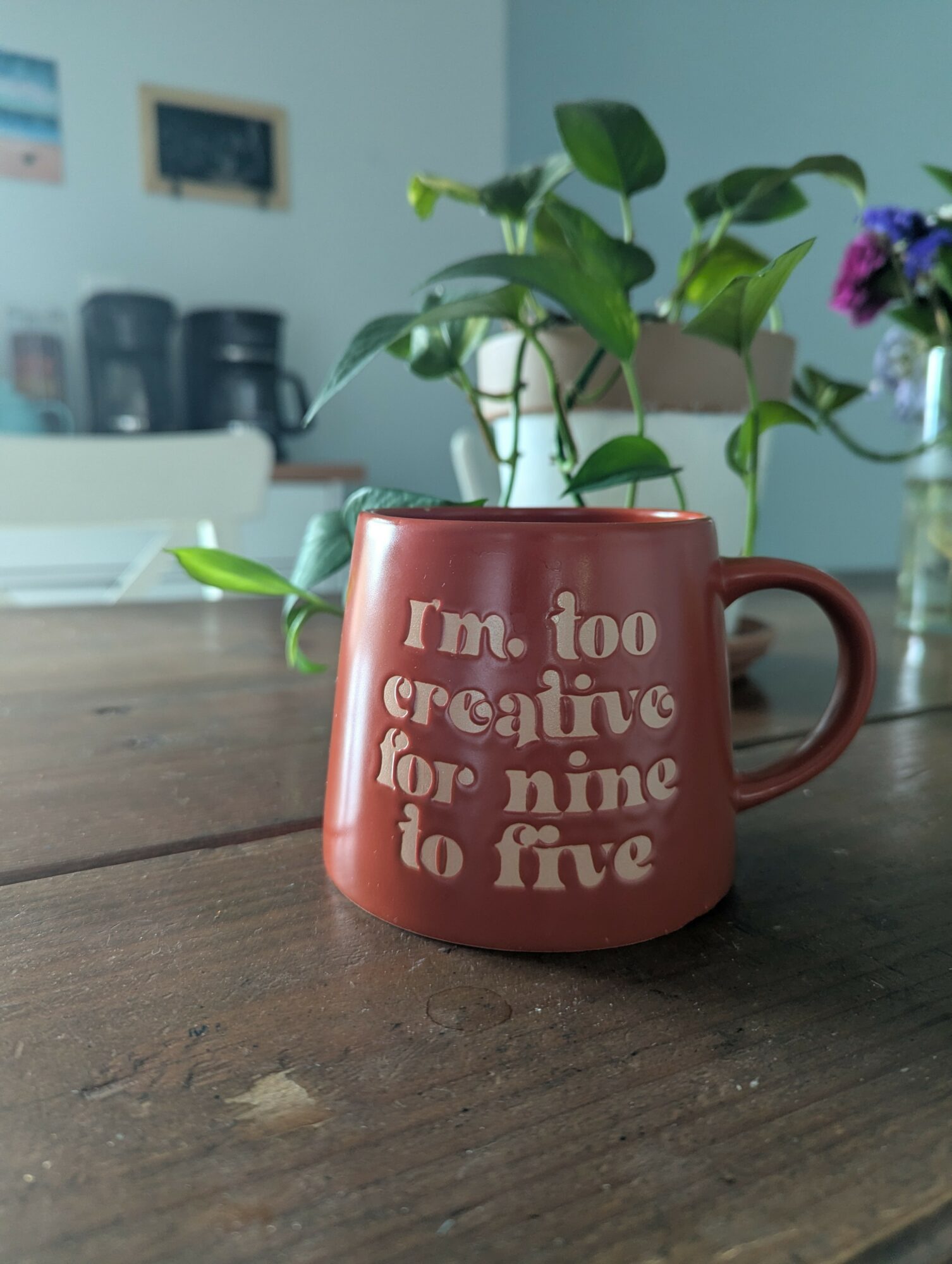 Red mug with white text on a wooden table, plant with green leaves in the background, and a blurred kitchen area.