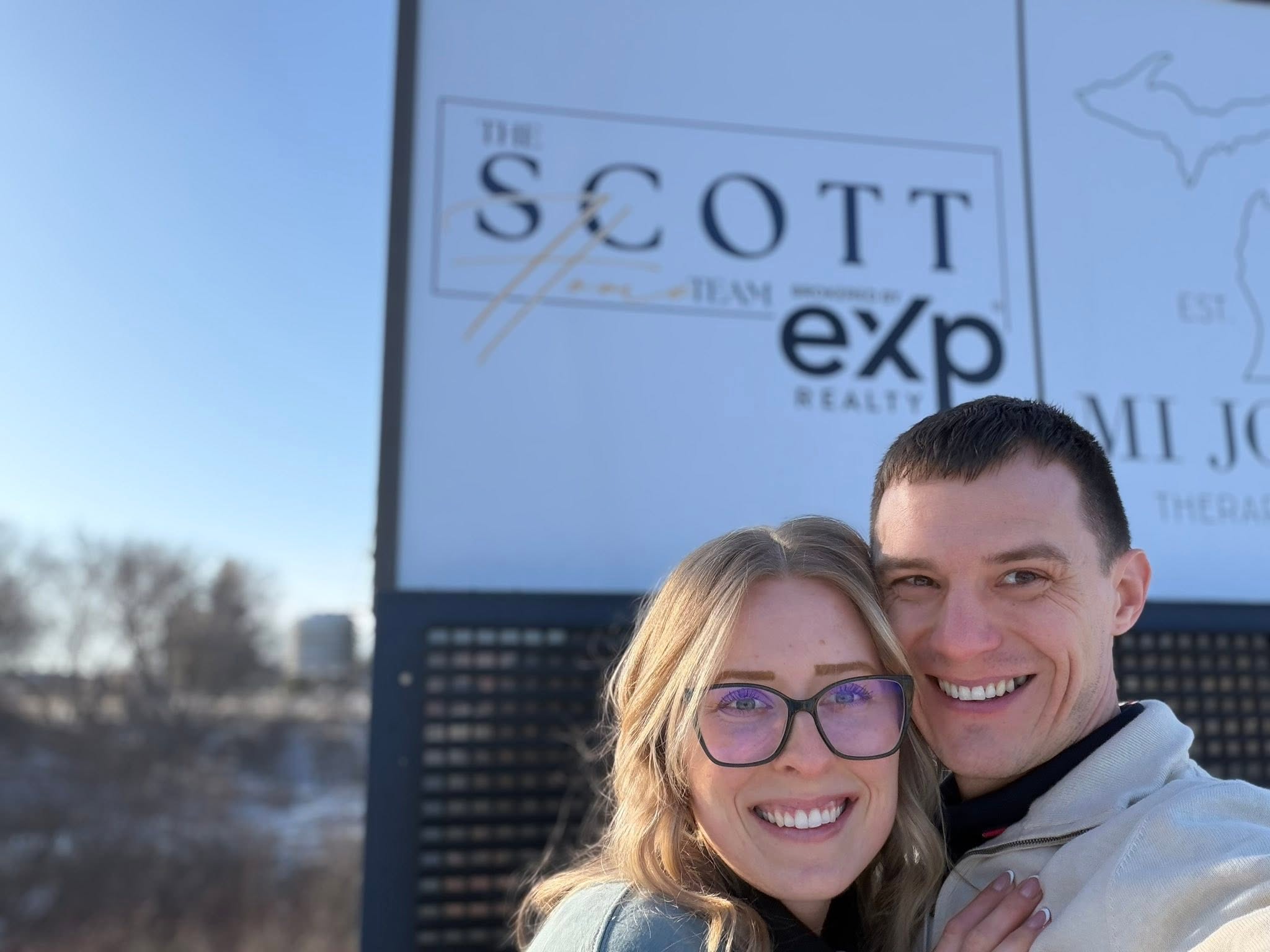 Two people smiling in front of a sign with text and a map, outdoors on a clear day.