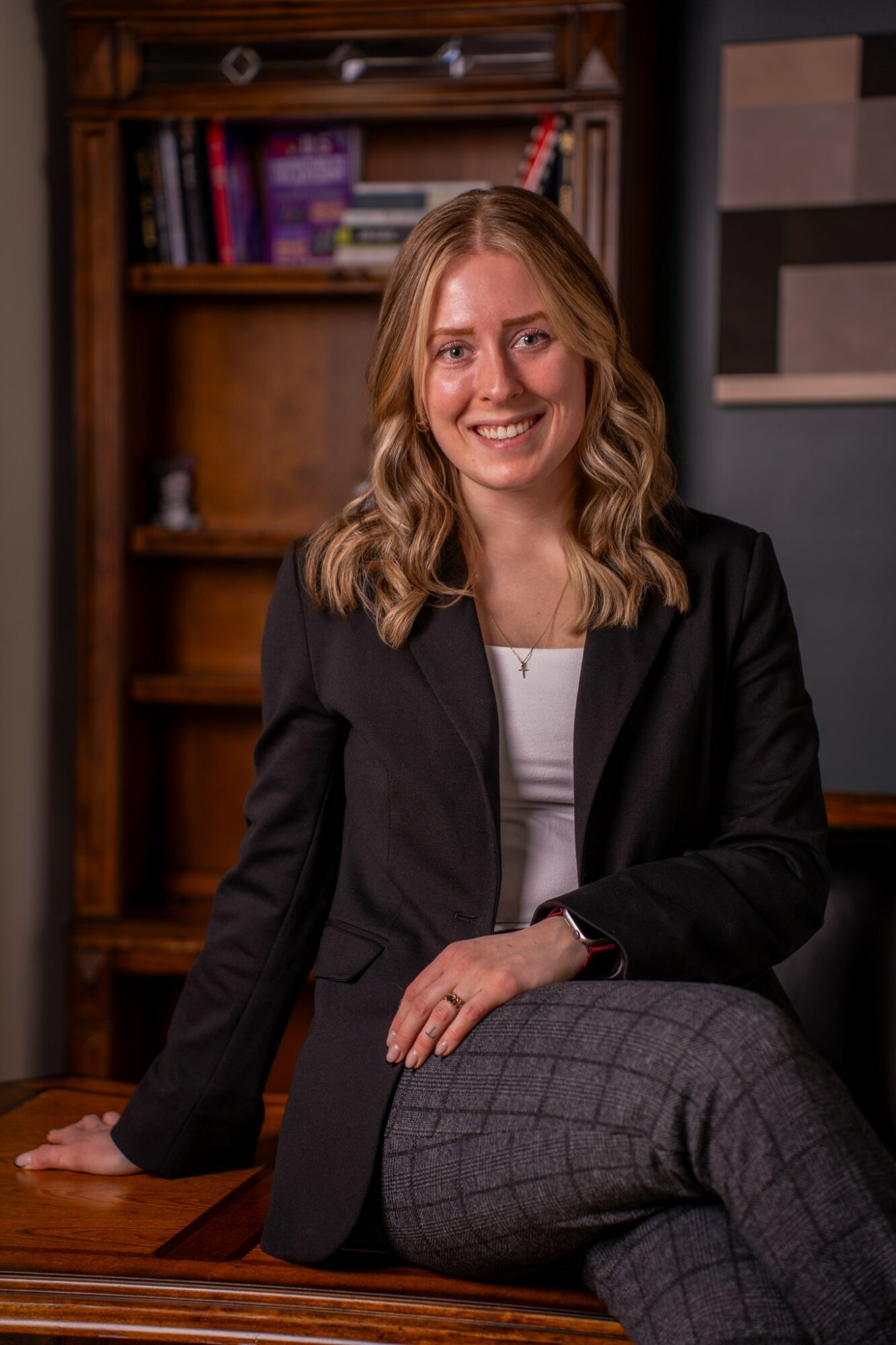 Woman with wavy blonde hair smiling, sitting on a wooden desk in a room with bookshelves, wearing a black blazer and gray checkered pants.