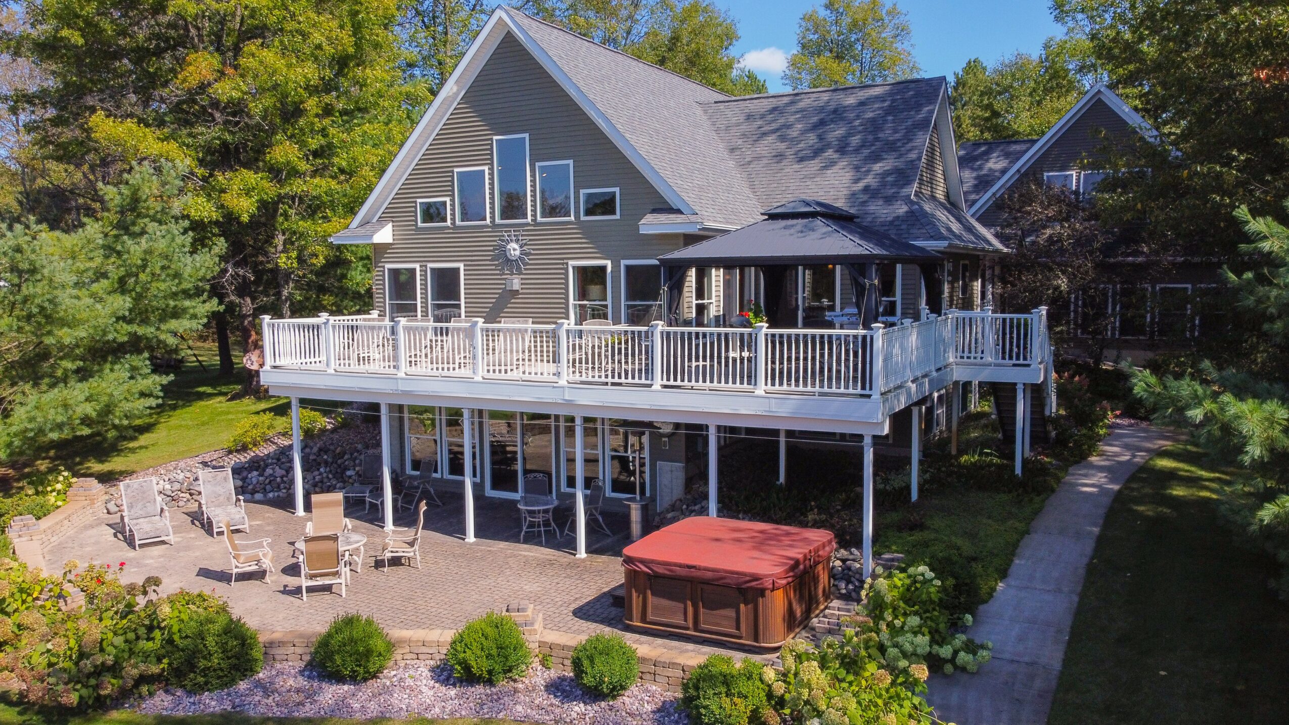 Large house with a spacious deck, surrounded by trees and a garden, with a pathway and outdoor furniture.