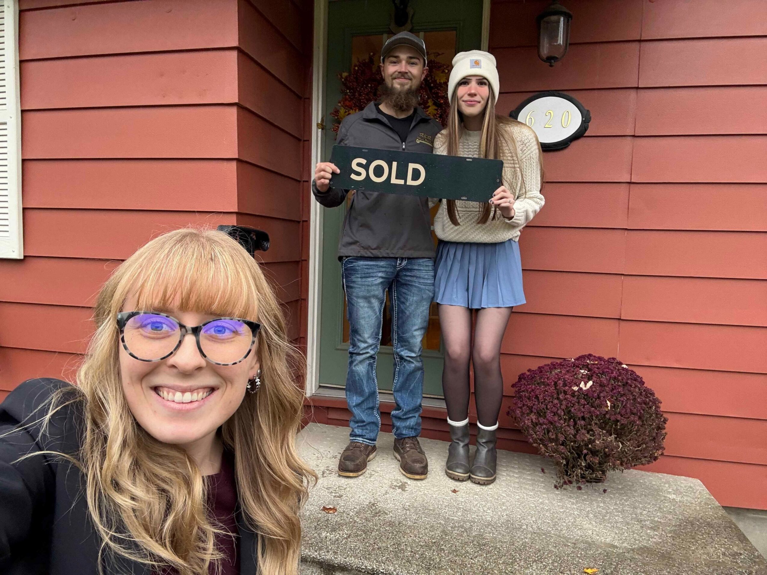 Two children and an adult woman stand outdoors on a porch, holding a sign that says 'SOLD'.
