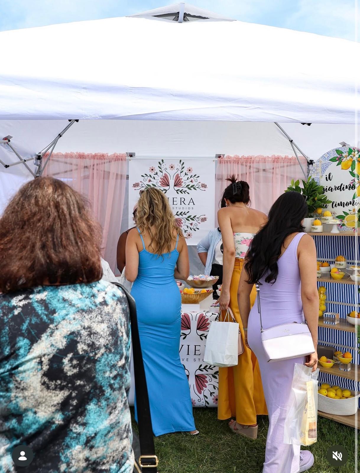 Four women stand under a white canopy at an outdoor event, with a display of lemons and a banner behind them.