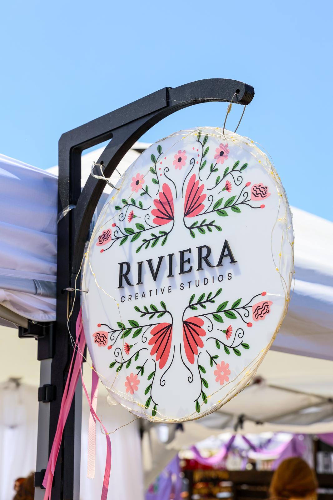 Round sign with floral design and pink butterflies, displaying 'RIVIERA CREATIVITY STUDIOS' text, hanging outdoors against a blue sky.