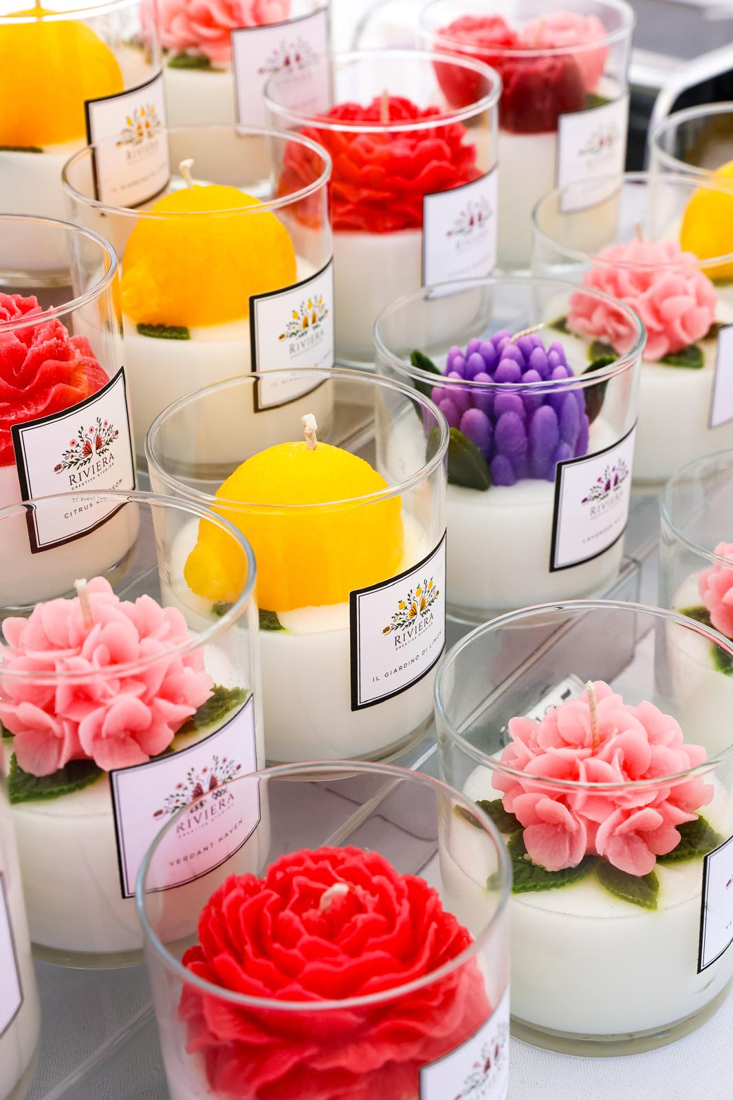 Colorful candles shaped like flowers and fruits in clear containers, with labels, arranged on a table.