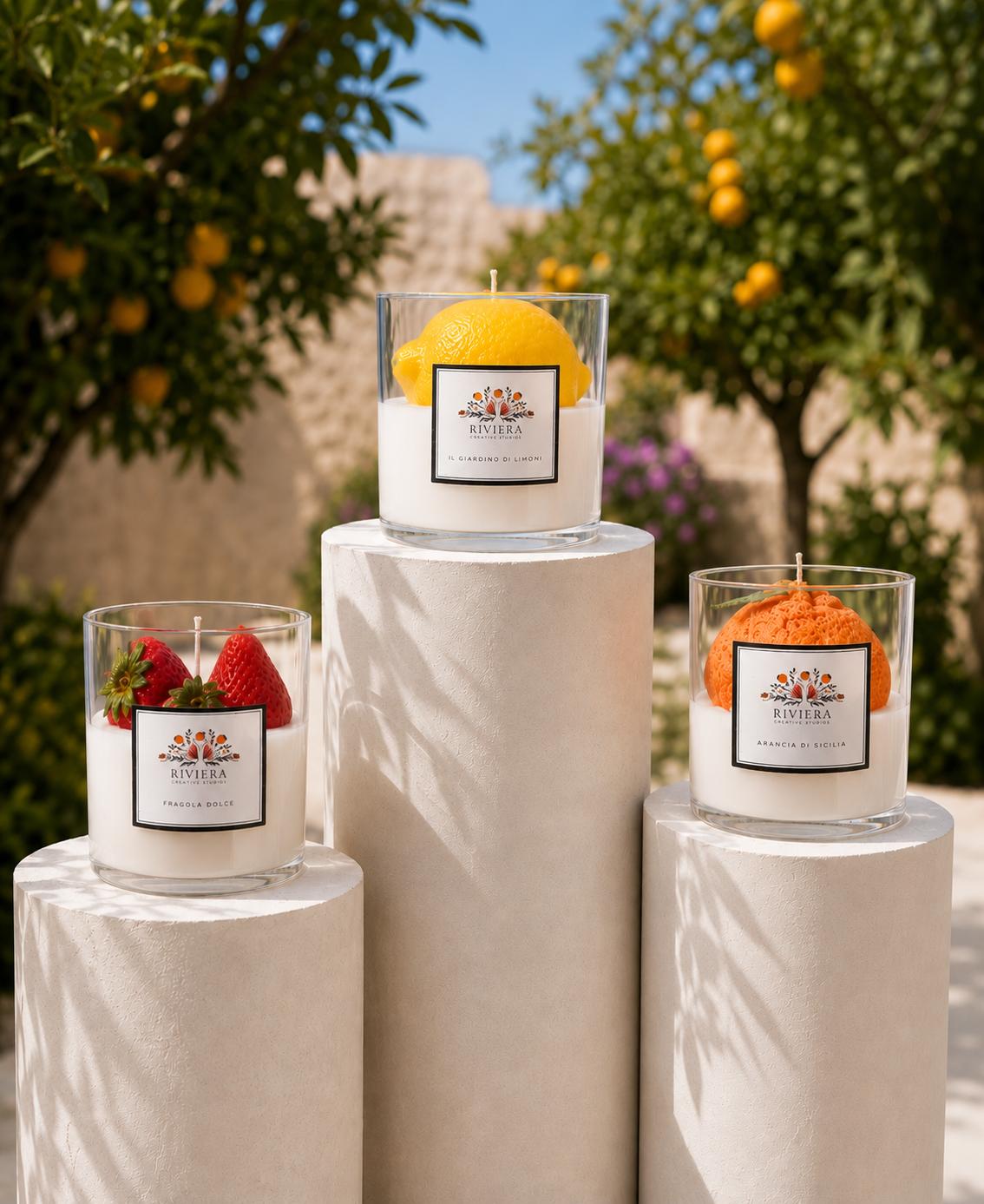 Three candles on white pedestals outdoors, with trees and yellow fruits in background, one with a lemon, others with strawberries and orange slices.