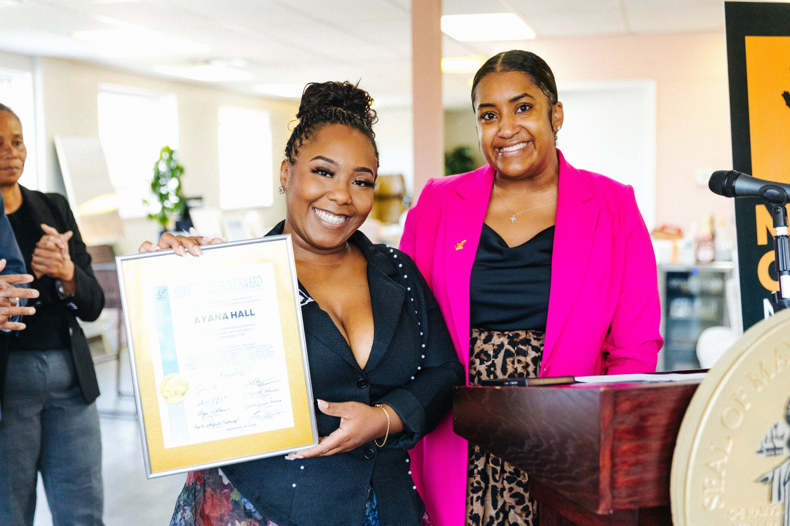 Two women smiling, one holding a framed certificate, in a bright indoor setting with other people in the background.