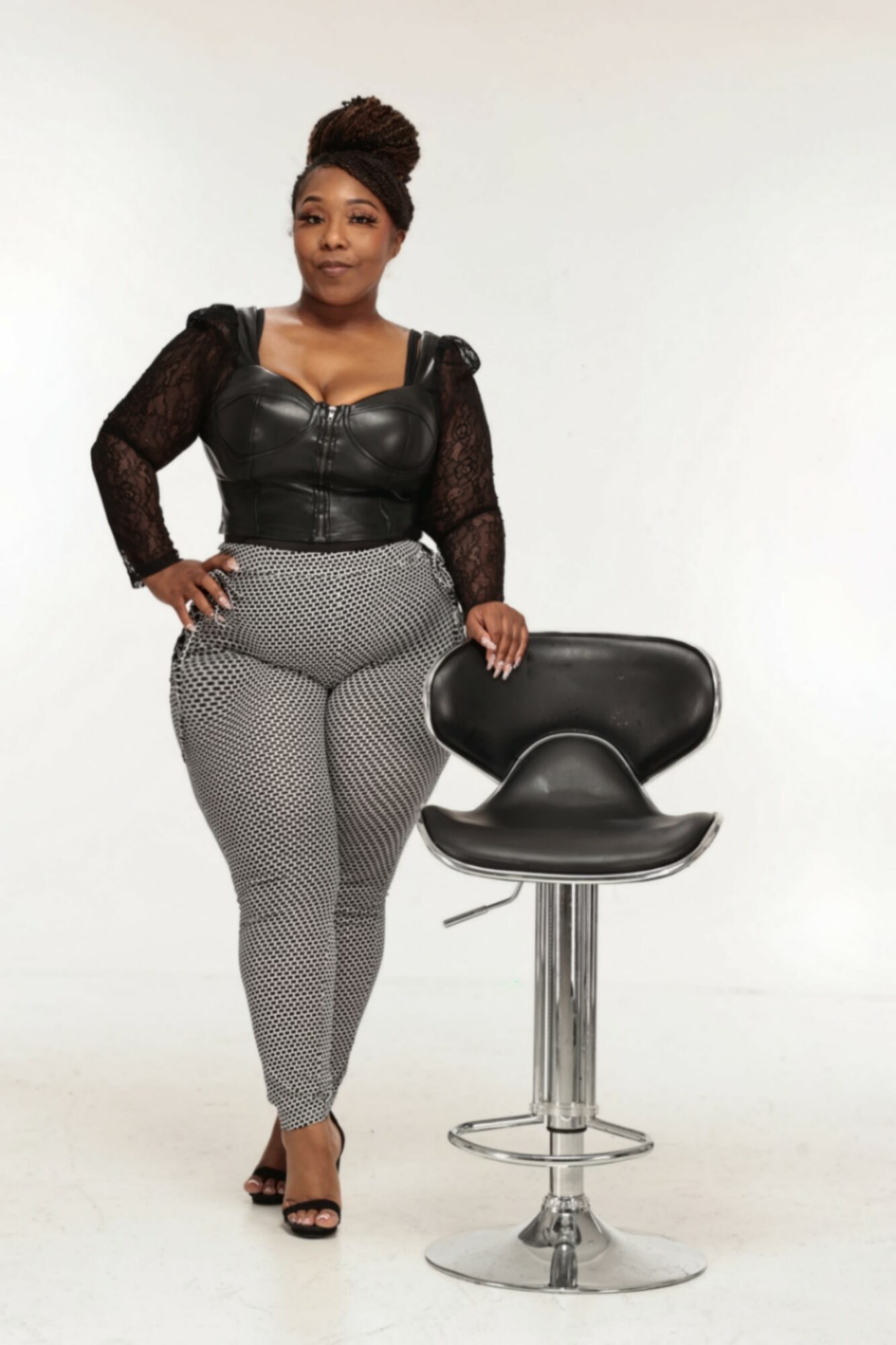Woman standing next to a black barstool, wearing a black top and patterned pants, with a neutral background.