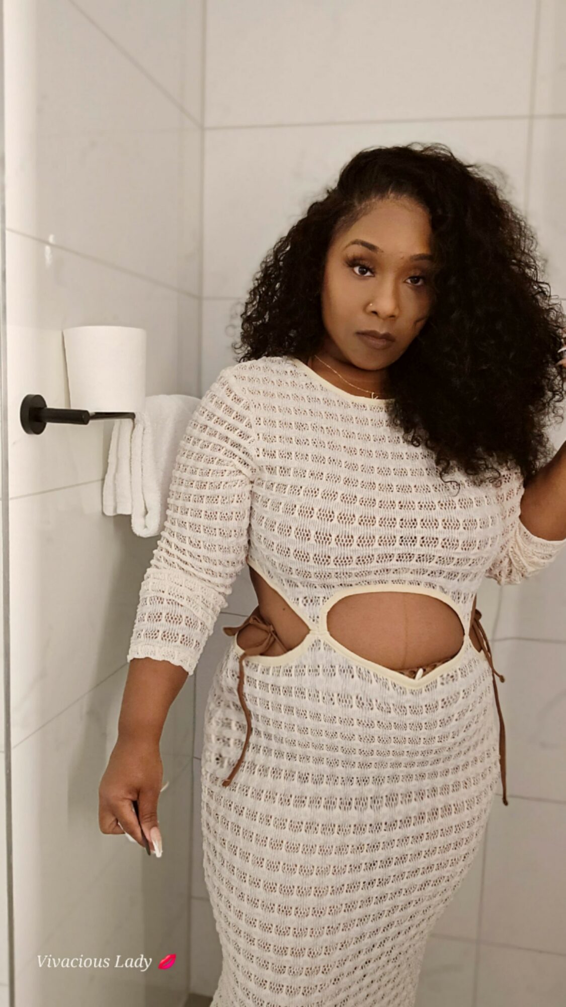Woman with curly hair in a white patterned dress with cutouts, standing in a bathroom with white tiles.