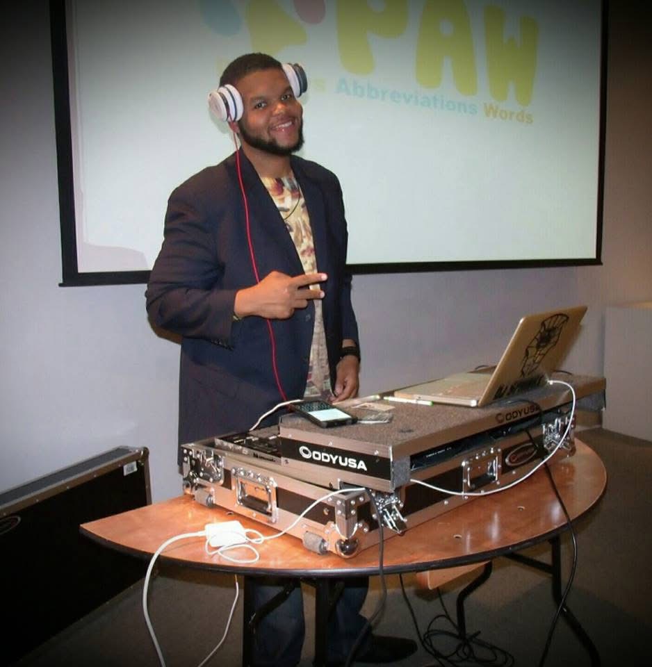 Man with headphones standing behind DJ equipment, smiling, in front of a screen with a logo. Table with laptop and DJ gear.
