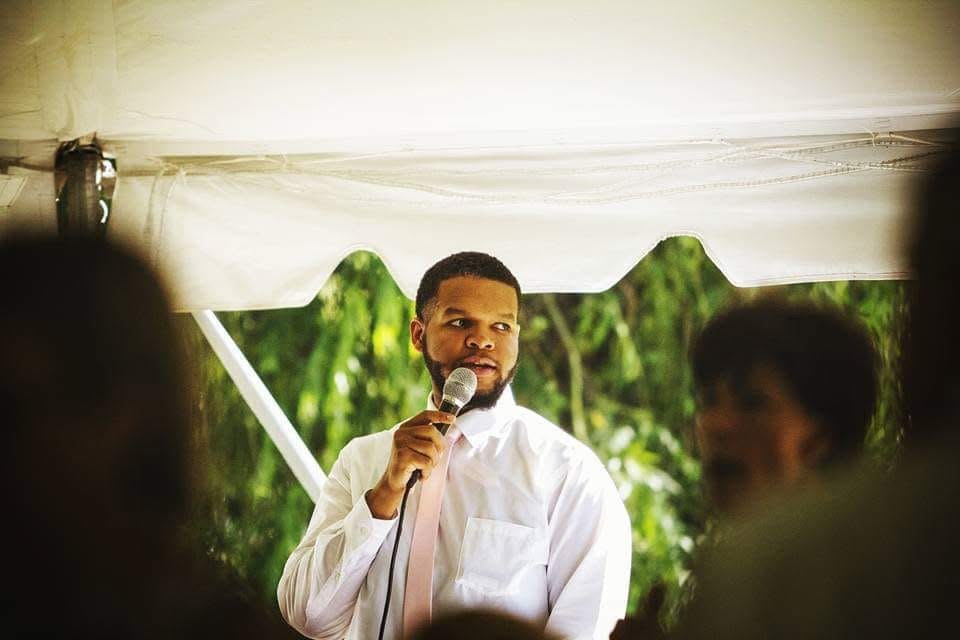 Man in white shirt holding a microphone, standing outdoors under a tent with green trees in the background.