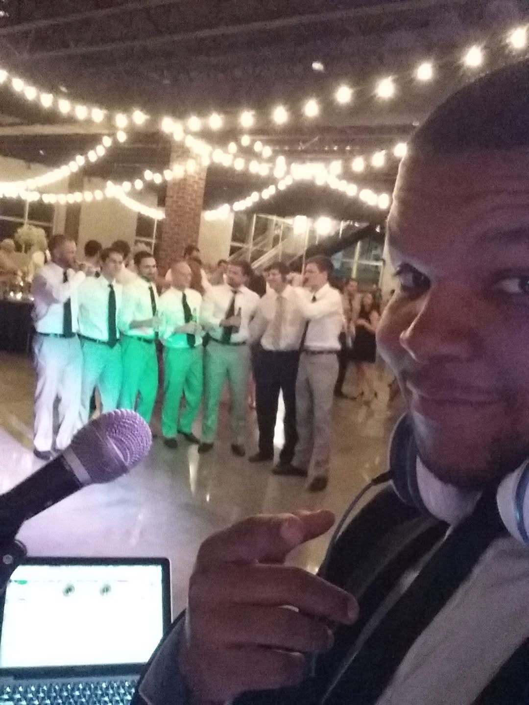 Person taking a selfie at a party with a group of people dancing in the background under string lights.