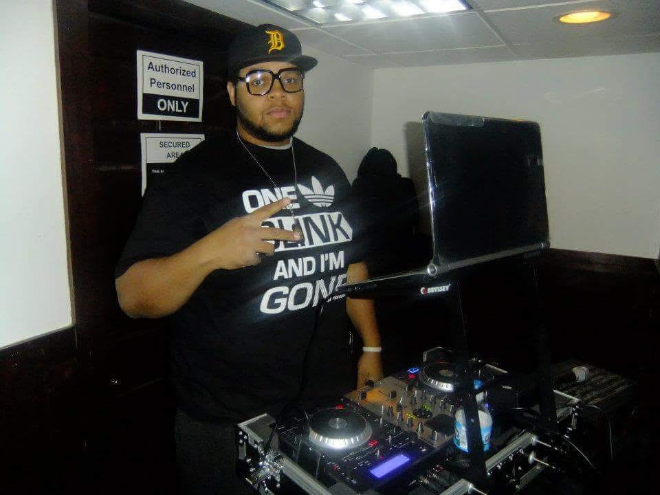 Person wearing glasses and a black cap stands next to DJ equipment, pointing at the camera, in a room with signs on the wall.