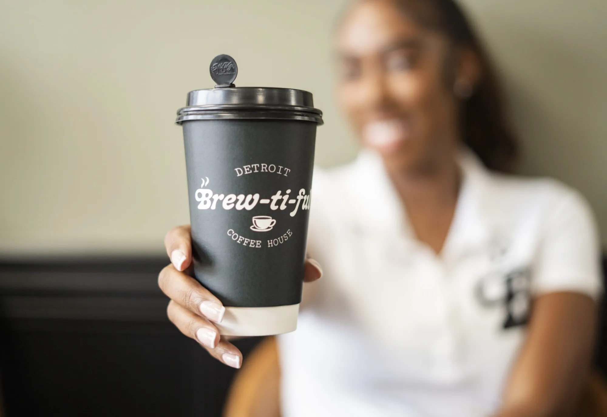 Person holding a black coffee cup with a logo, blurred background, person wearing white shirt, smiling.