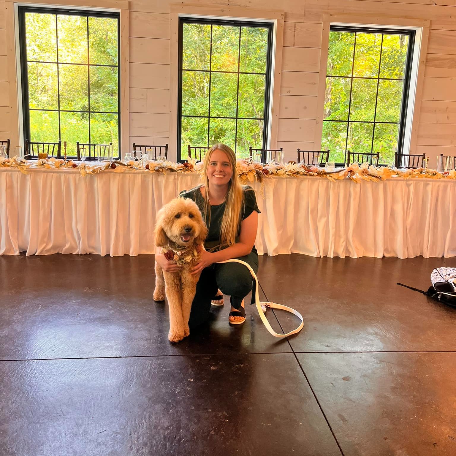 Woman kneeling with a fluffy dog in front of a long table with pink tablecloths, windows showing green trees outside.